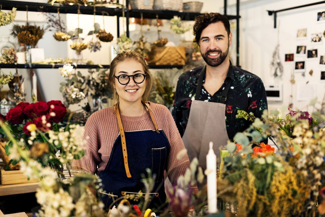 Two people in aprons smiling, surrounded by flowers in a flower shop.