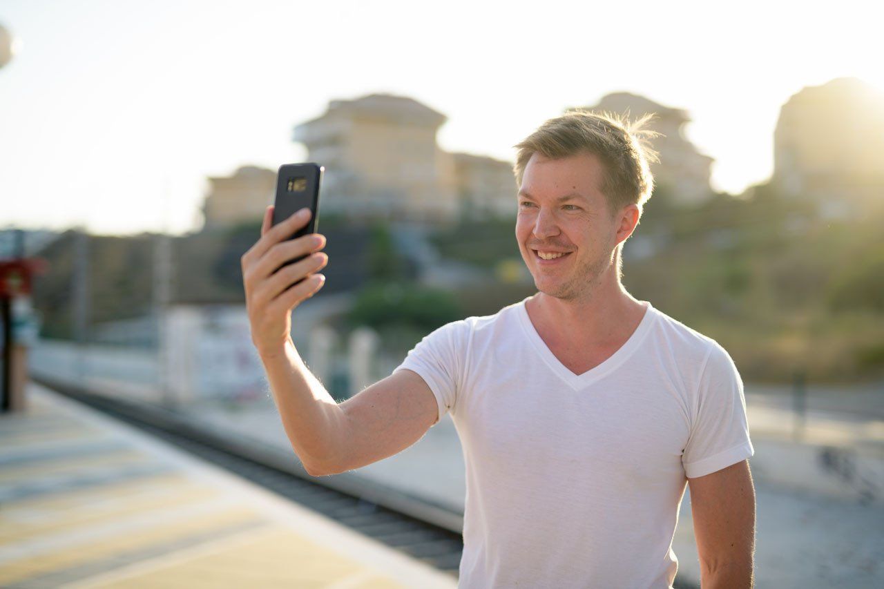 Man in white shirt takes a selfie with a phone, smiling outdoors in sunlight.