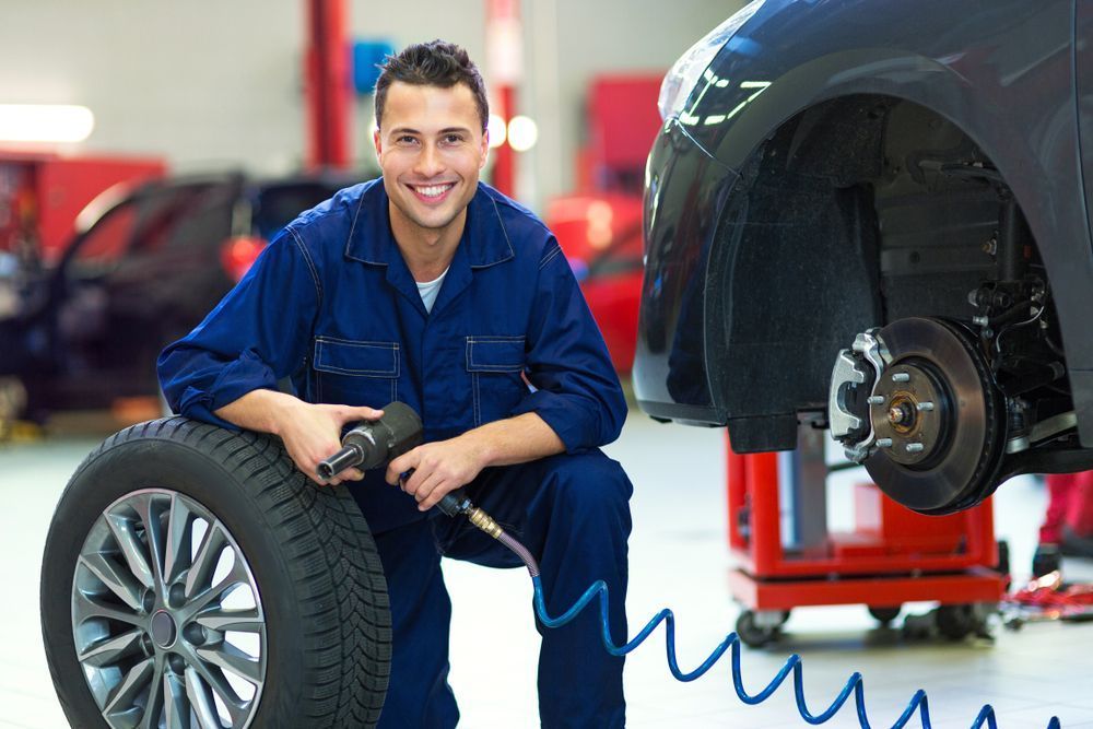 Mechanic smiling, holding tire and impact wrench, working on a car in a garage.