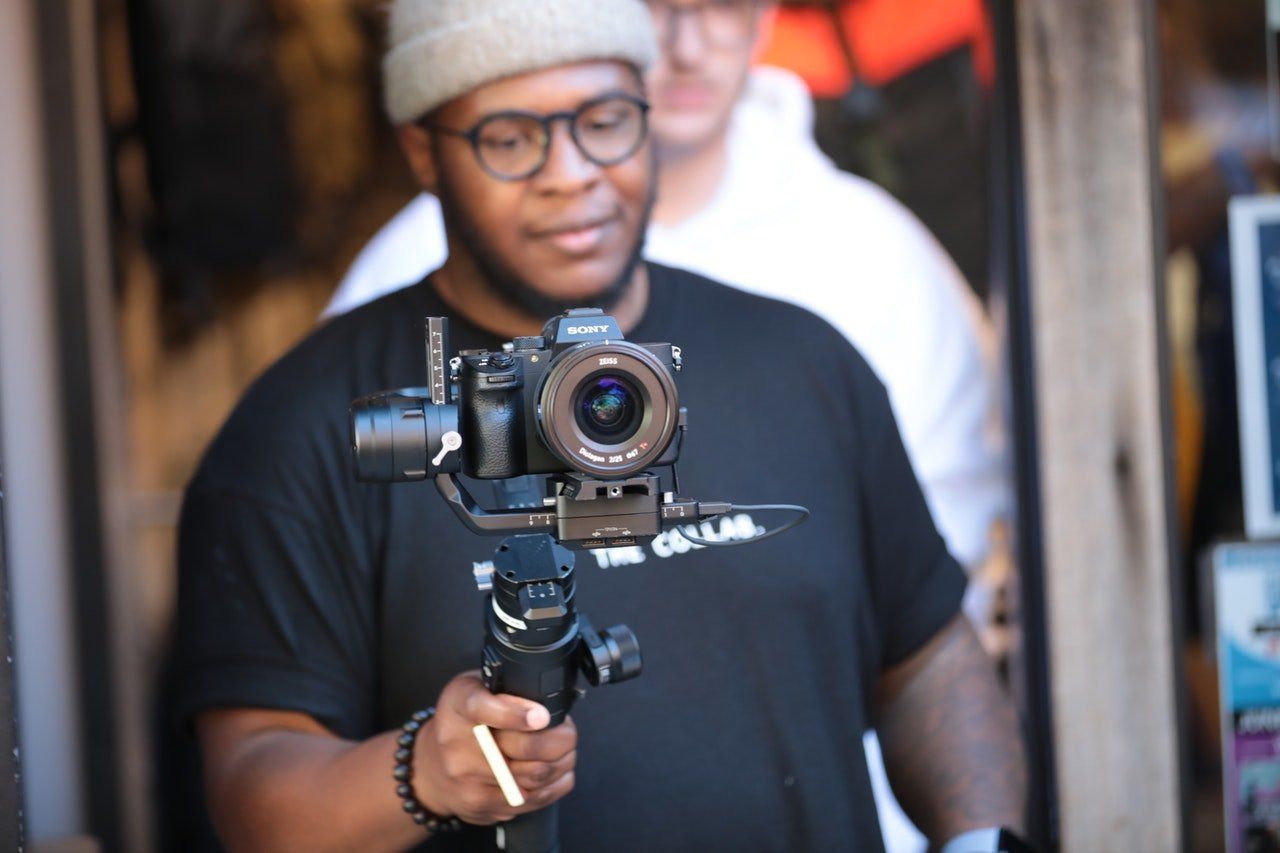 Man holding a camera on a stabilizer in an open doorway. He wears glasses, a black shirt, and a beanie.