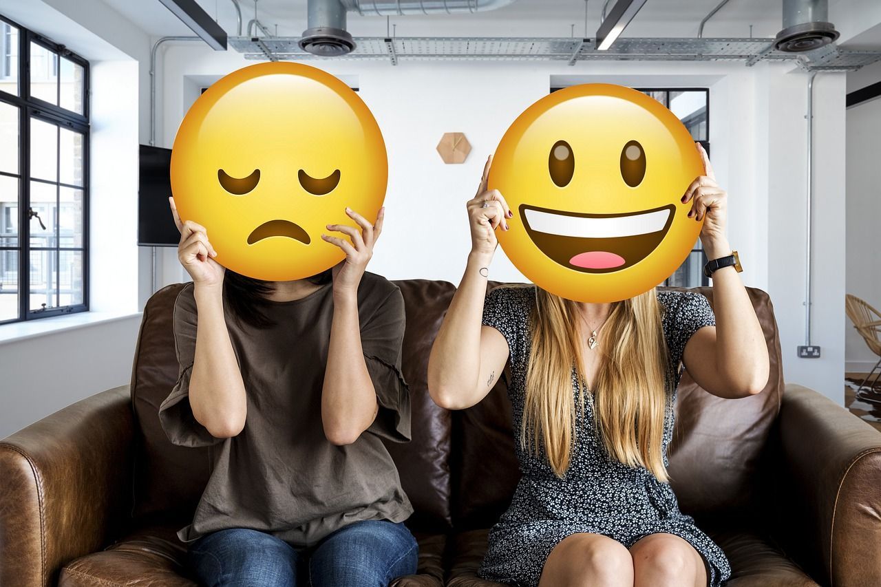 Two people on a couch hold up emoji masks: one sad, one happy.