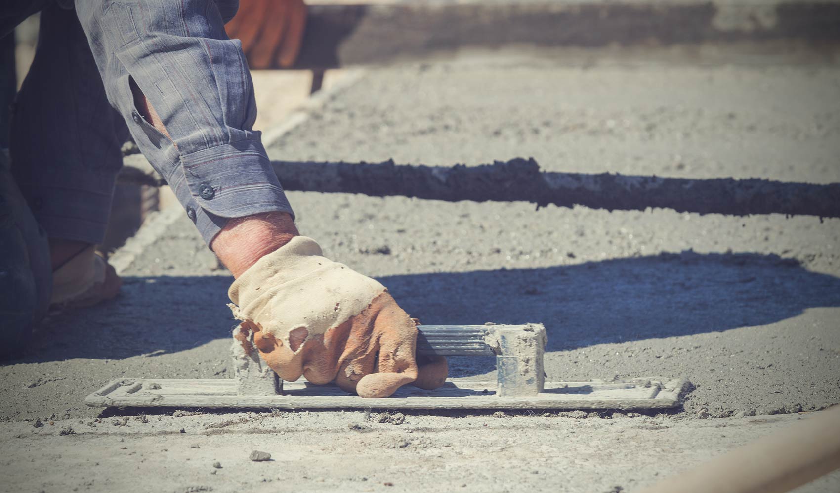 Construction worker wearing gloves smoothing wet concrete with a hand float tool.