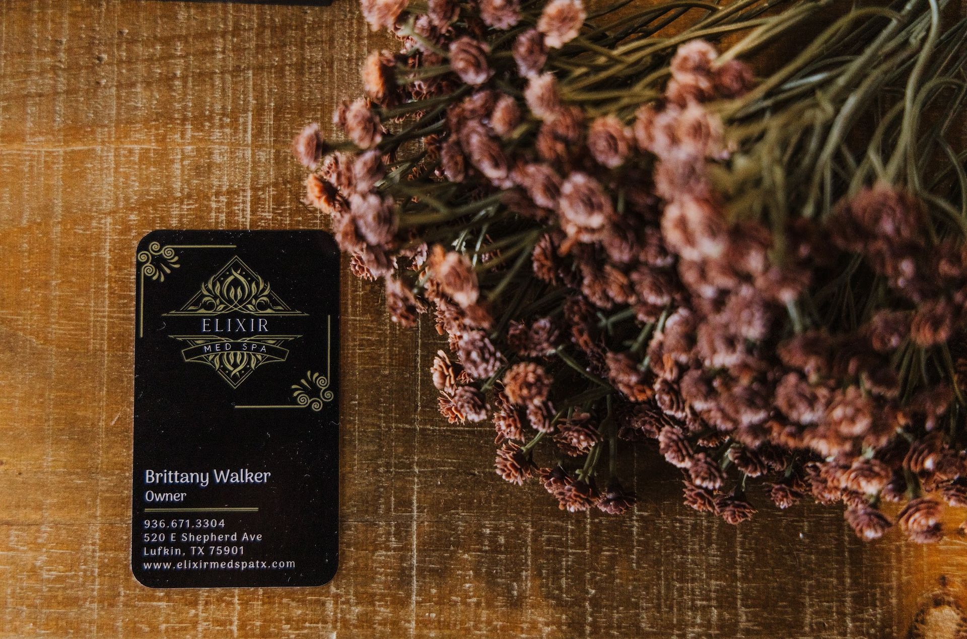 A business card is sitting on a wooden table next to a bunch of flowers.