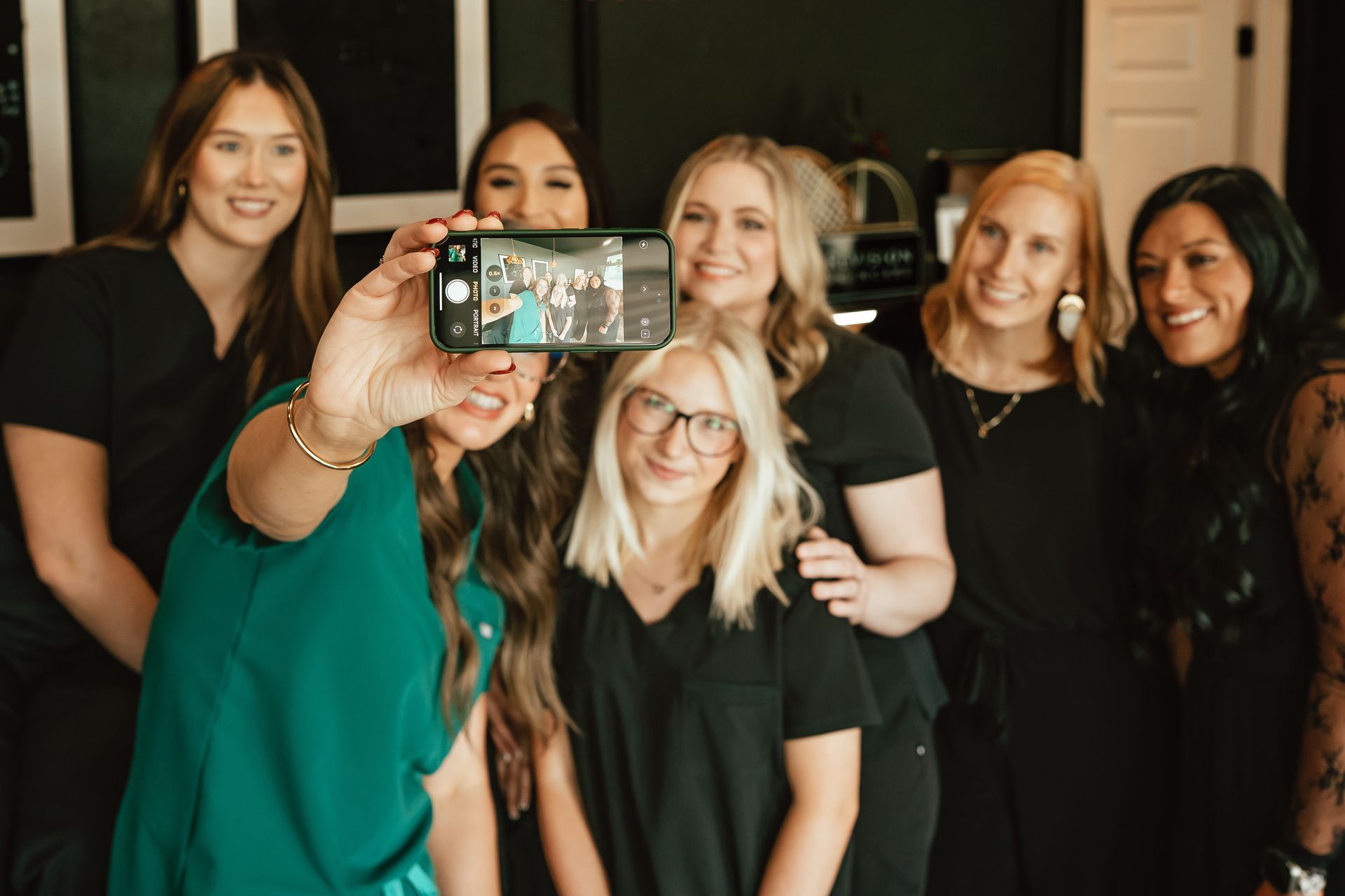 A group of seven women taking a selfie; dressed in scrubs, smiling, and in a room.
