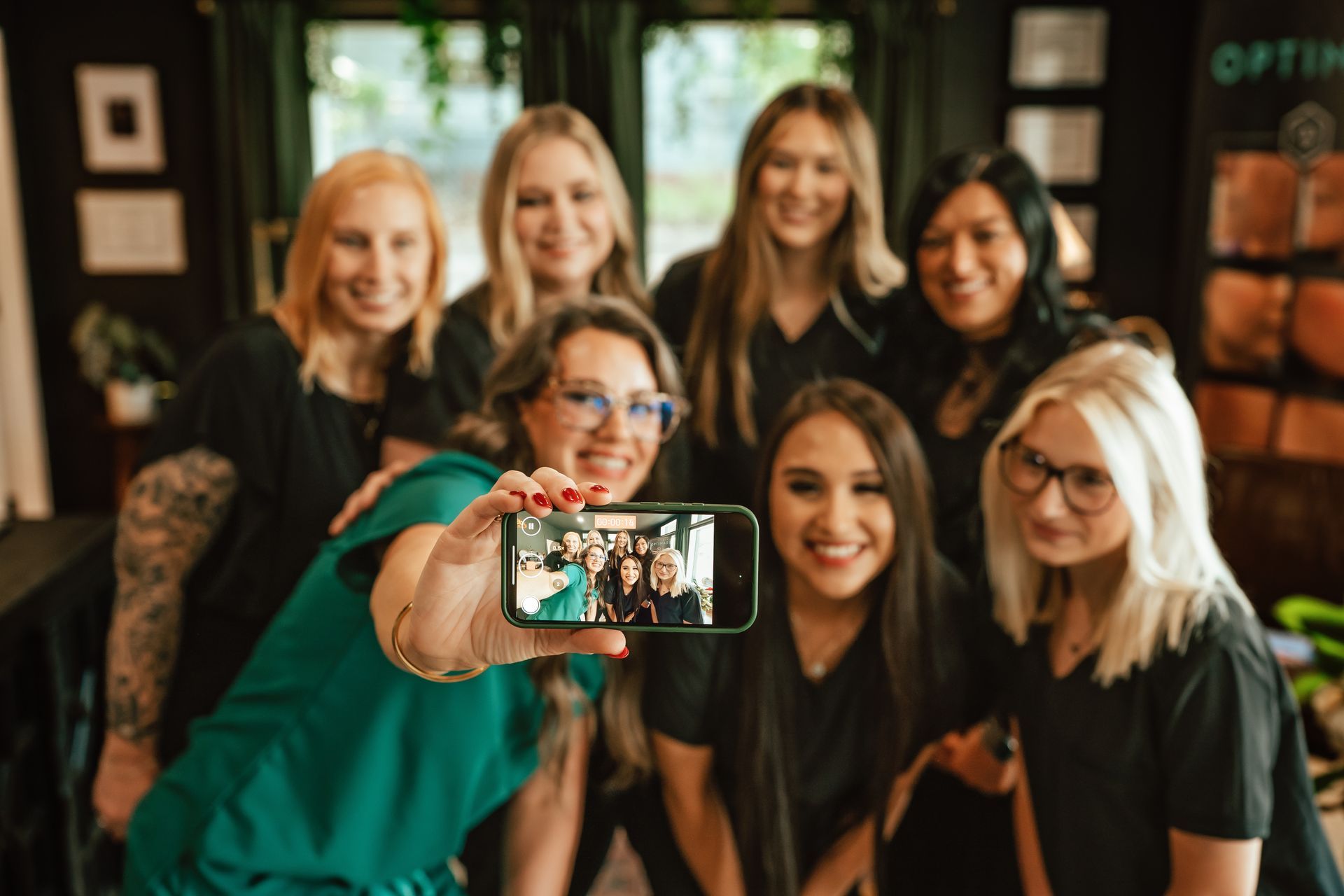 Group of seven women taking a selfie, smiling. They are in a dark room, some wearing glasses and dark tops.