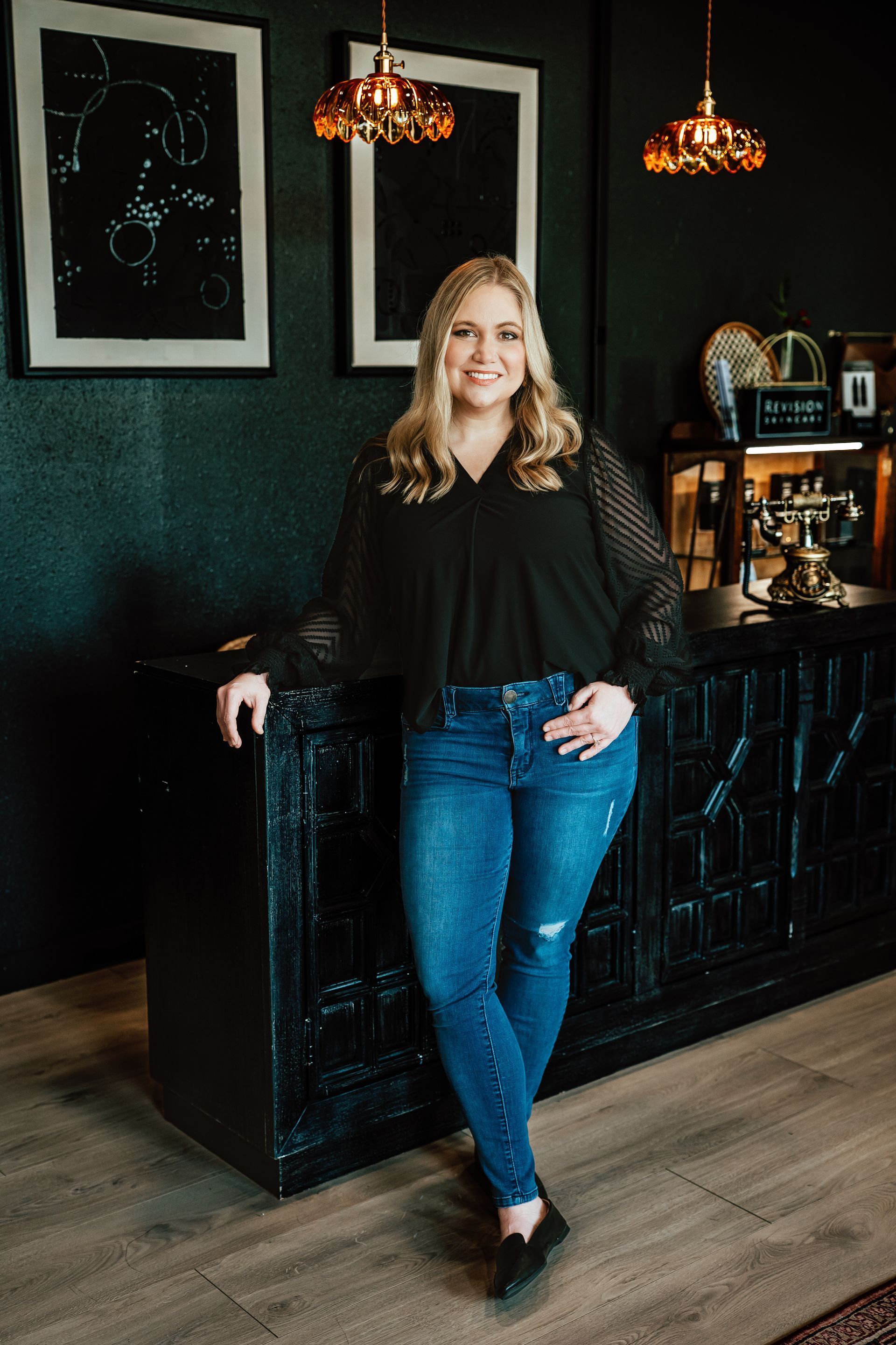 Woman in black top and jeans leaning on a dark bar, smiling.