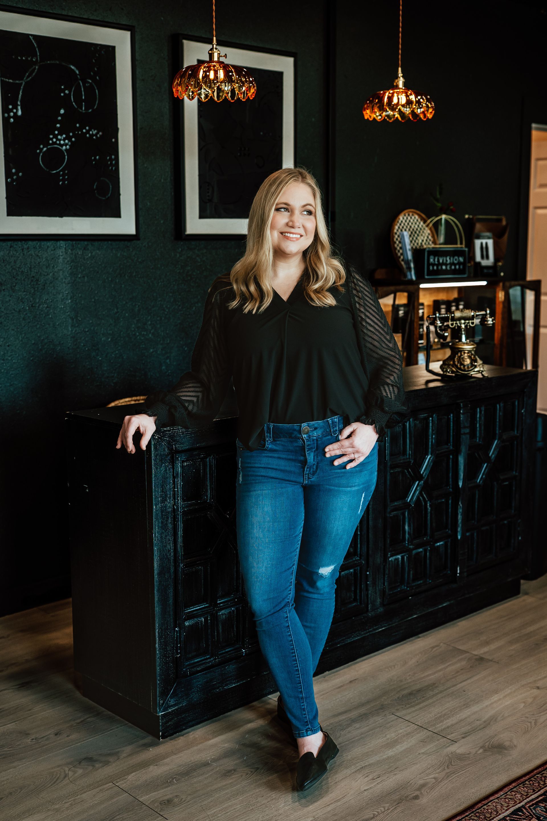 Woman in black top and jeans, leaning on a dark cabinet, smiling.