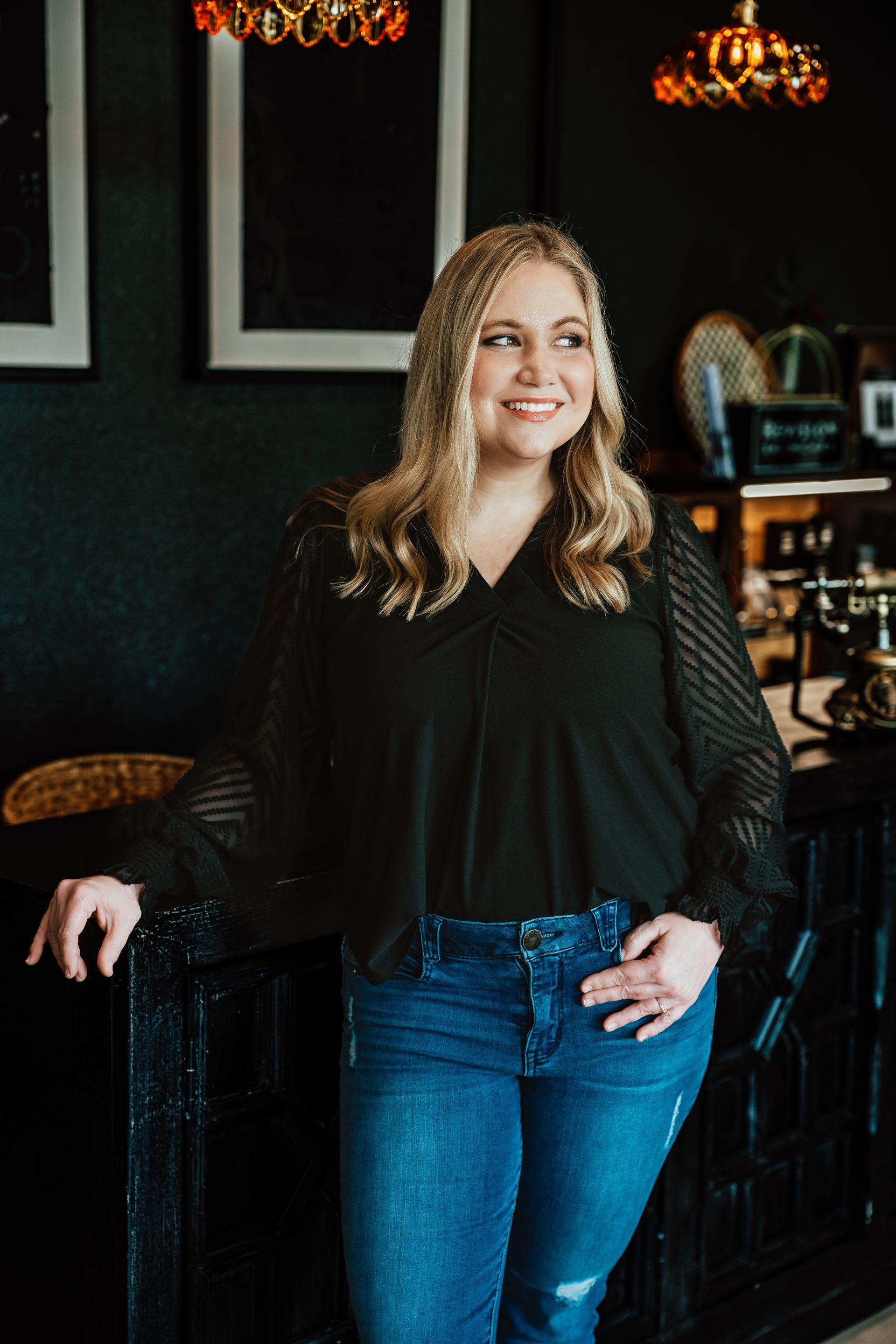 Woman with blonde hair, smiling, wearing a black top and jeans, leaning on a dark cabinet in a room.
