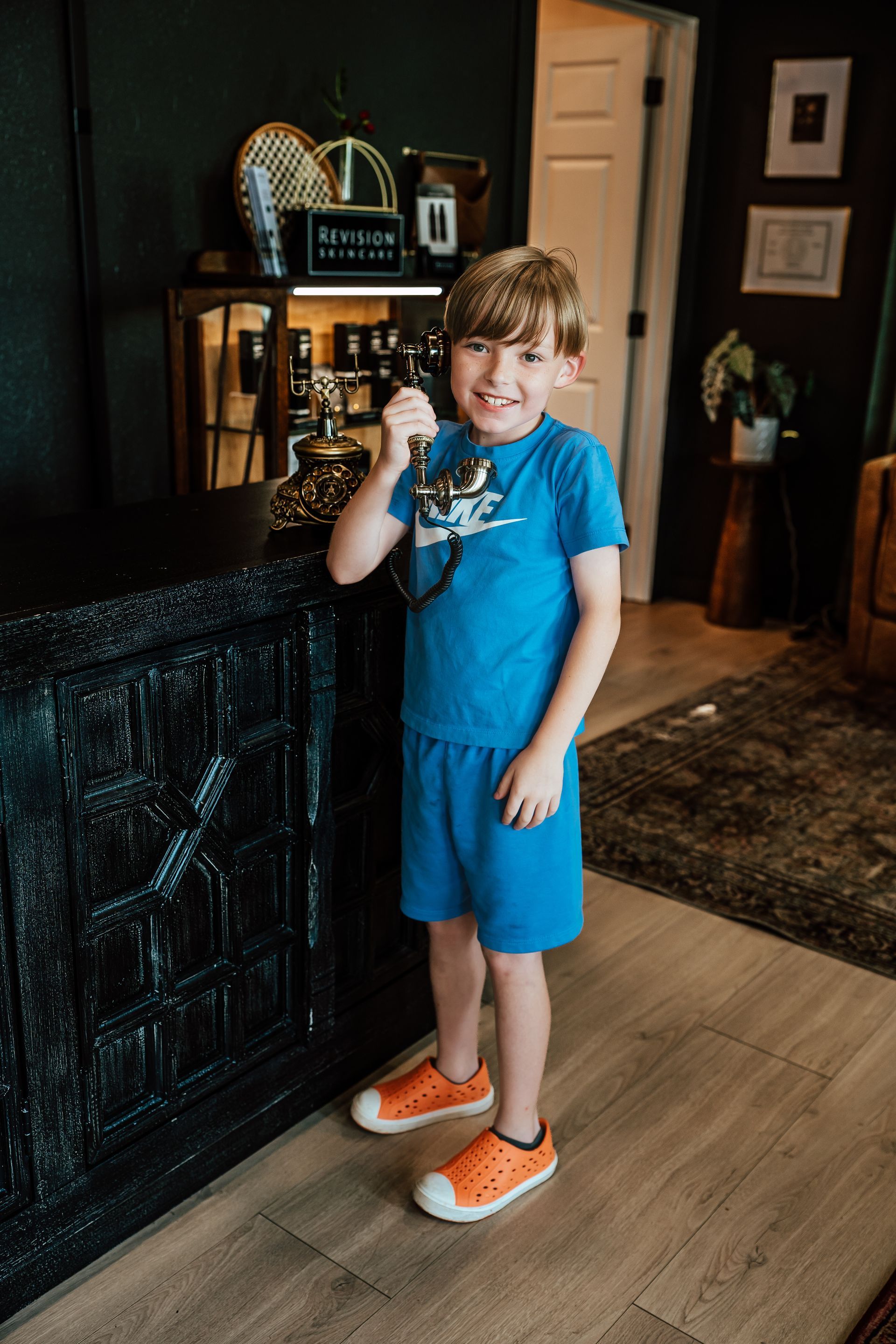 Boy in blue shirt and shorts, holding a vintage telephone, smiling in a room.