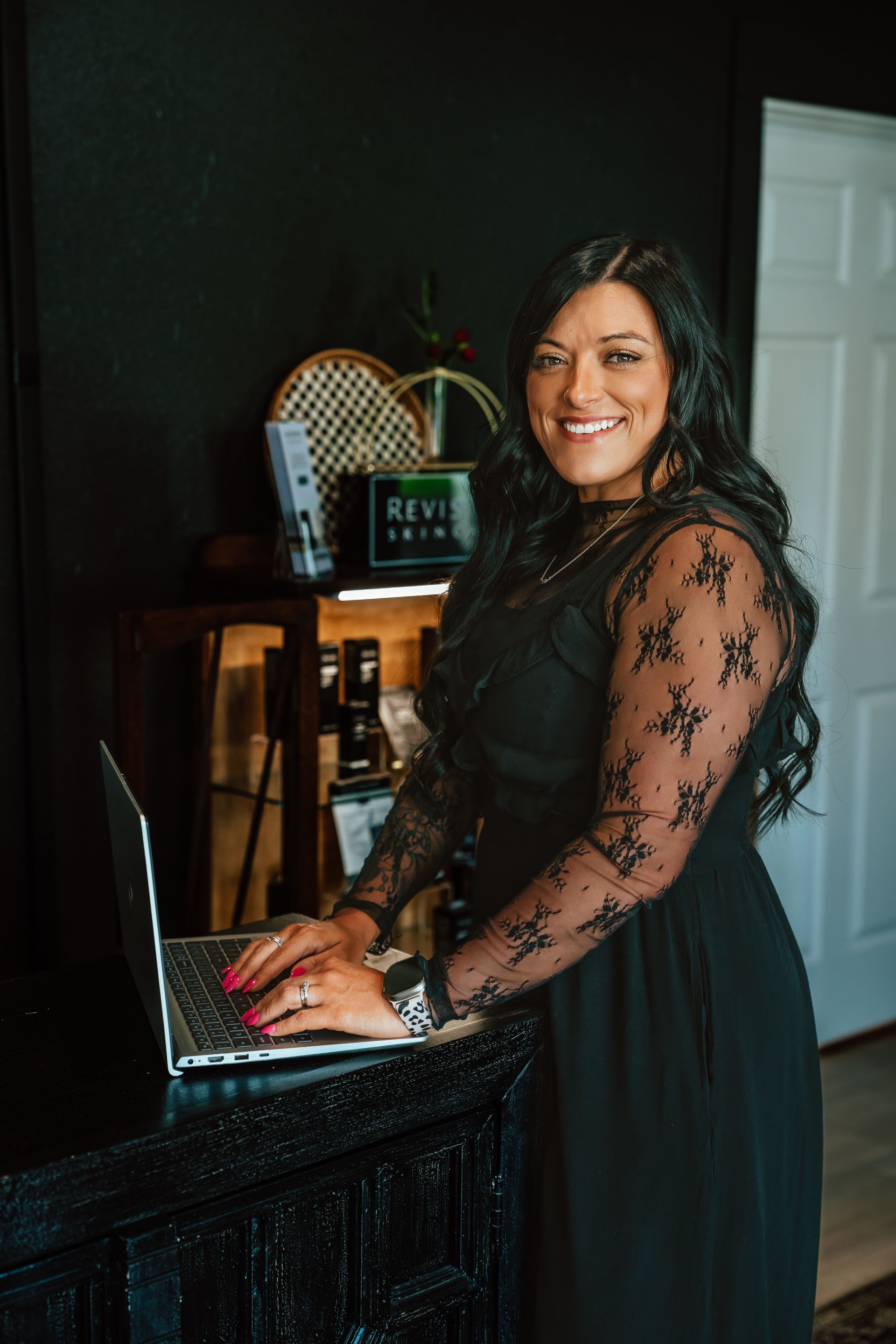 Woman in black dress, smiling, typing on laptop at dark counter; office setting.
