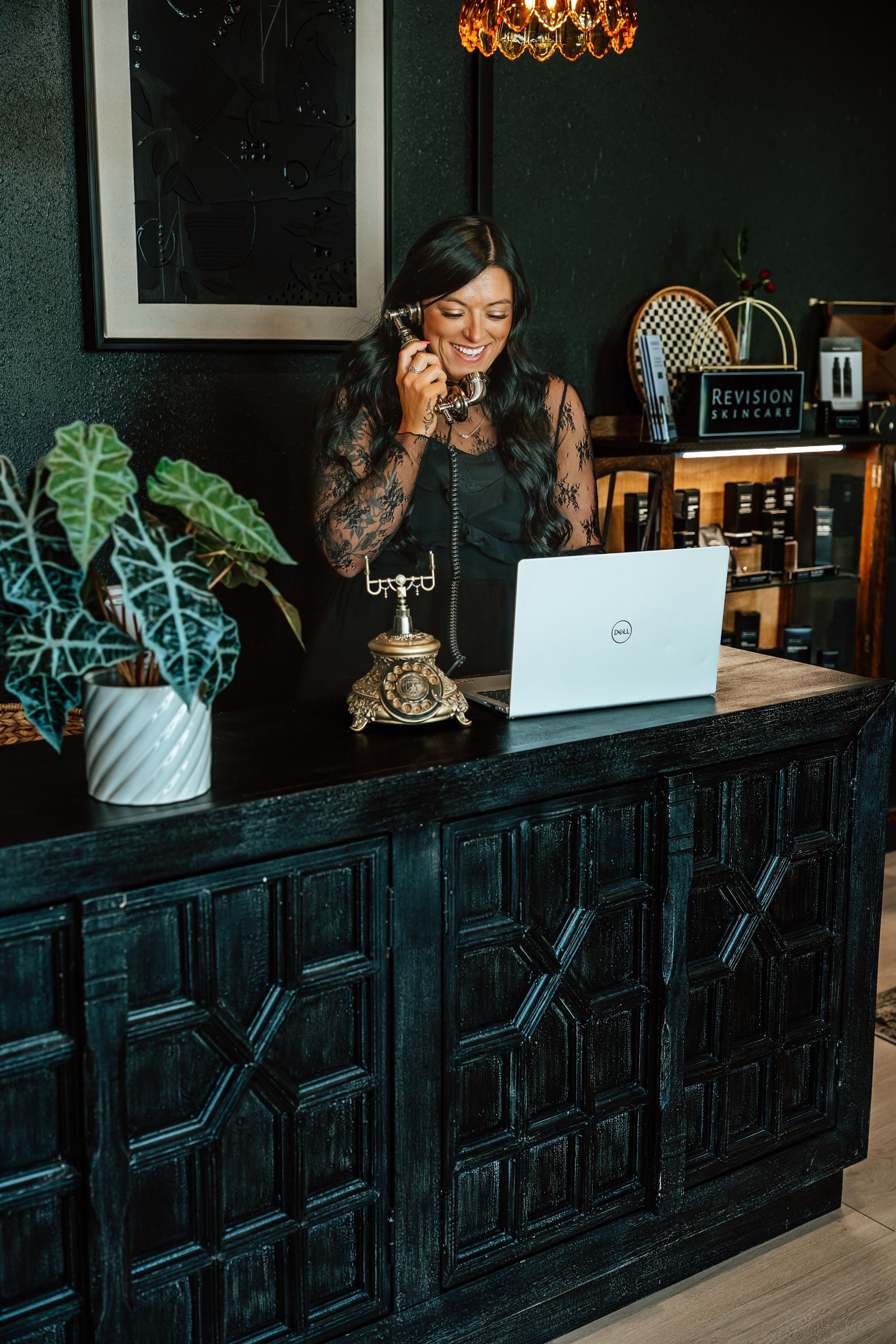 Woman in black lace top smiling, talking on a vintage phone at a dark reception desk.