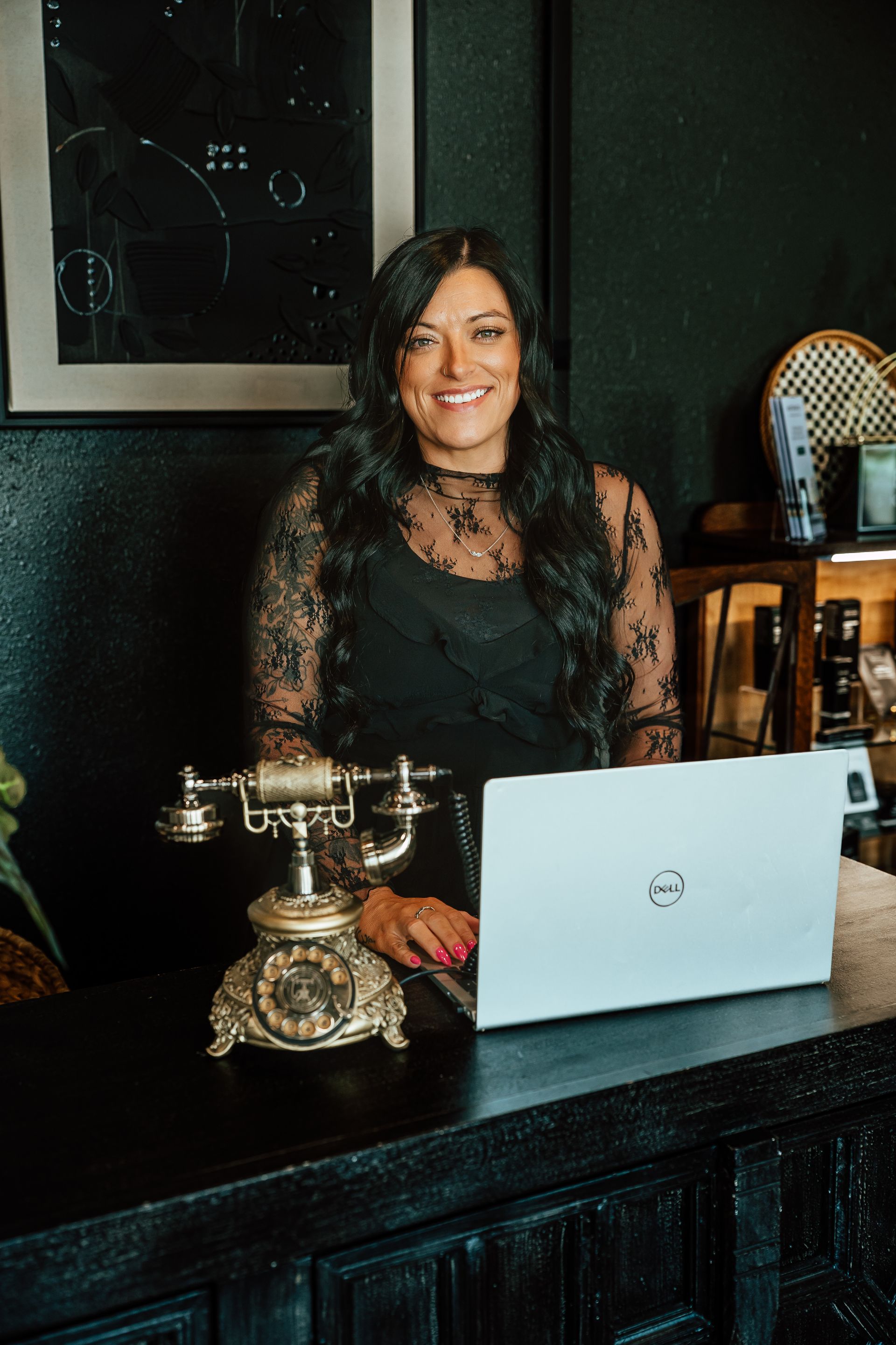 Woman with long dark hair smiles, working on laptop behind a desk, with vintage phone. Dark room.