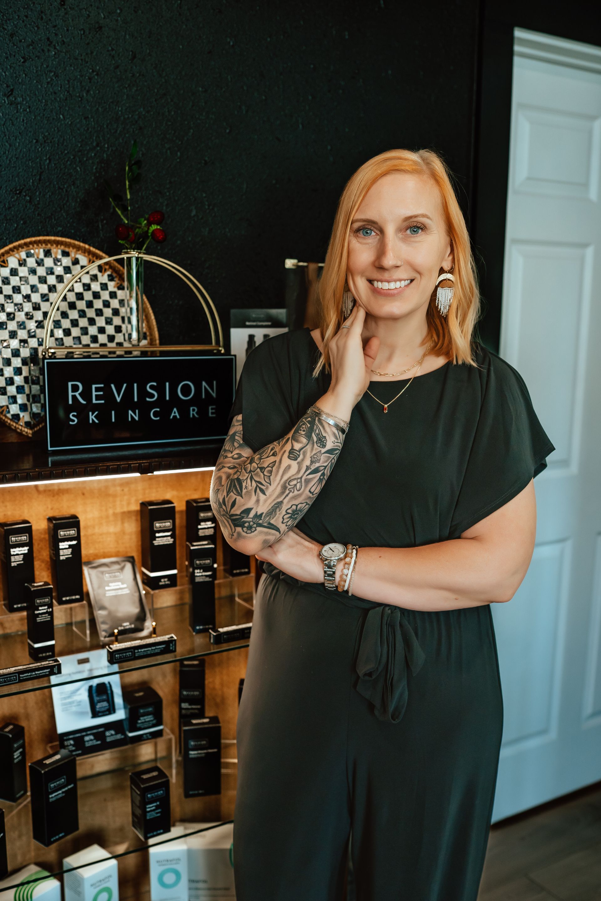 Woman with blonde hair, arm tattoos, in a skincare shop, posing by Revision Skincare products.