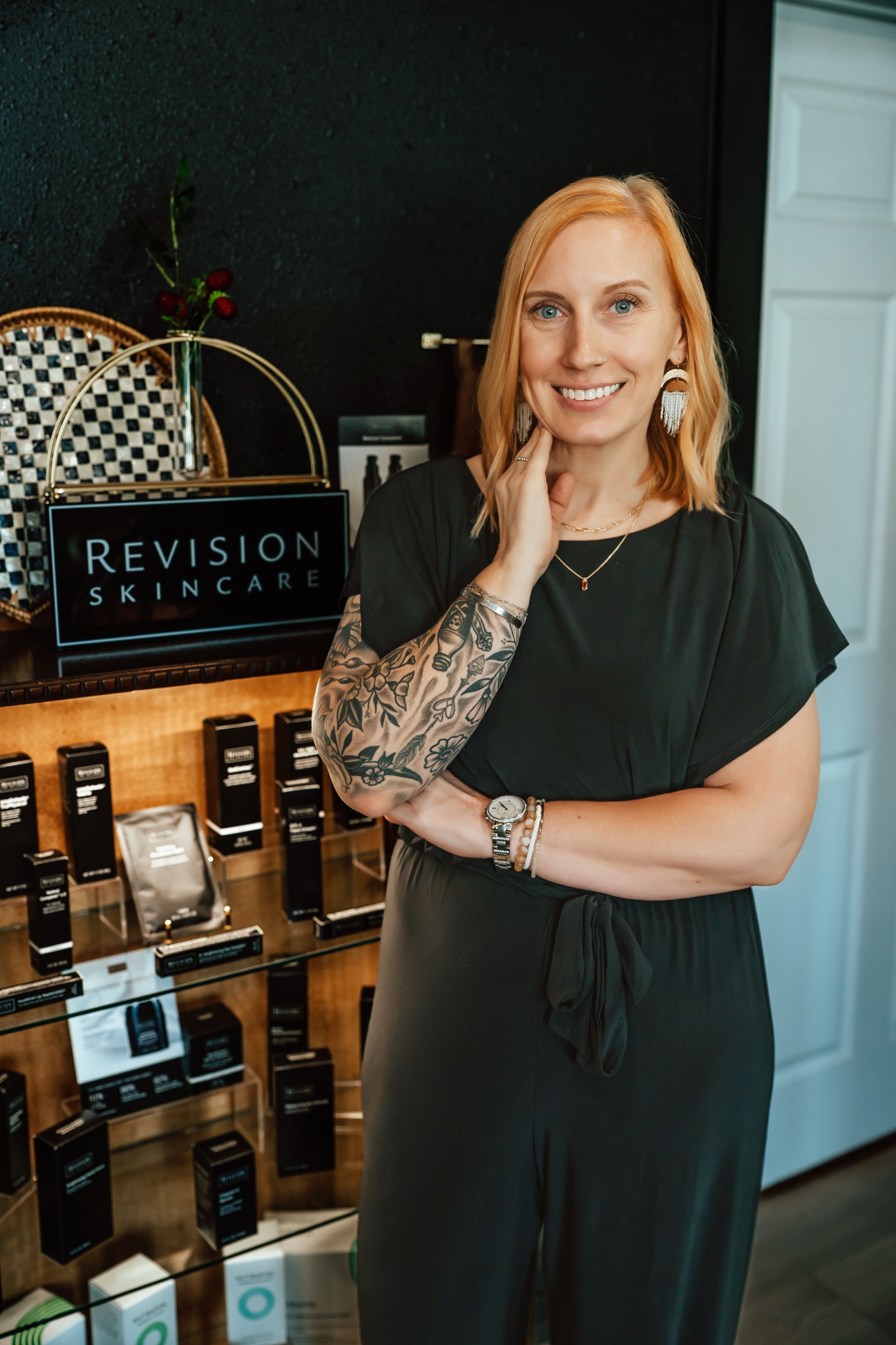 Woman in a skincare shop, smiles.  Arm tattoo, short blonde hair, products displayed with “Revision Skincare” sign.