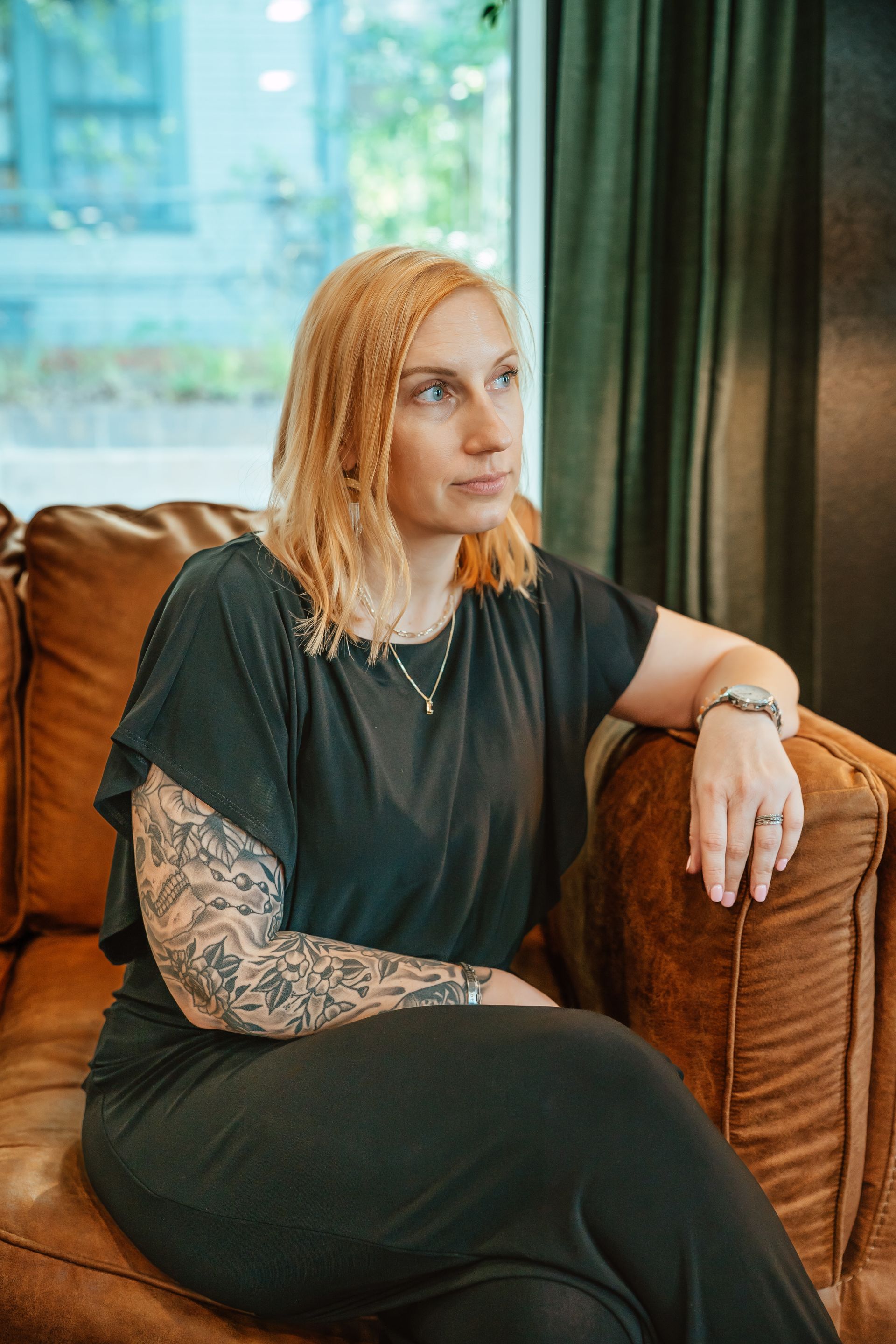 Blonde woman with tattoos on a brown leather couch, looking thoughtful; indoors, green background.