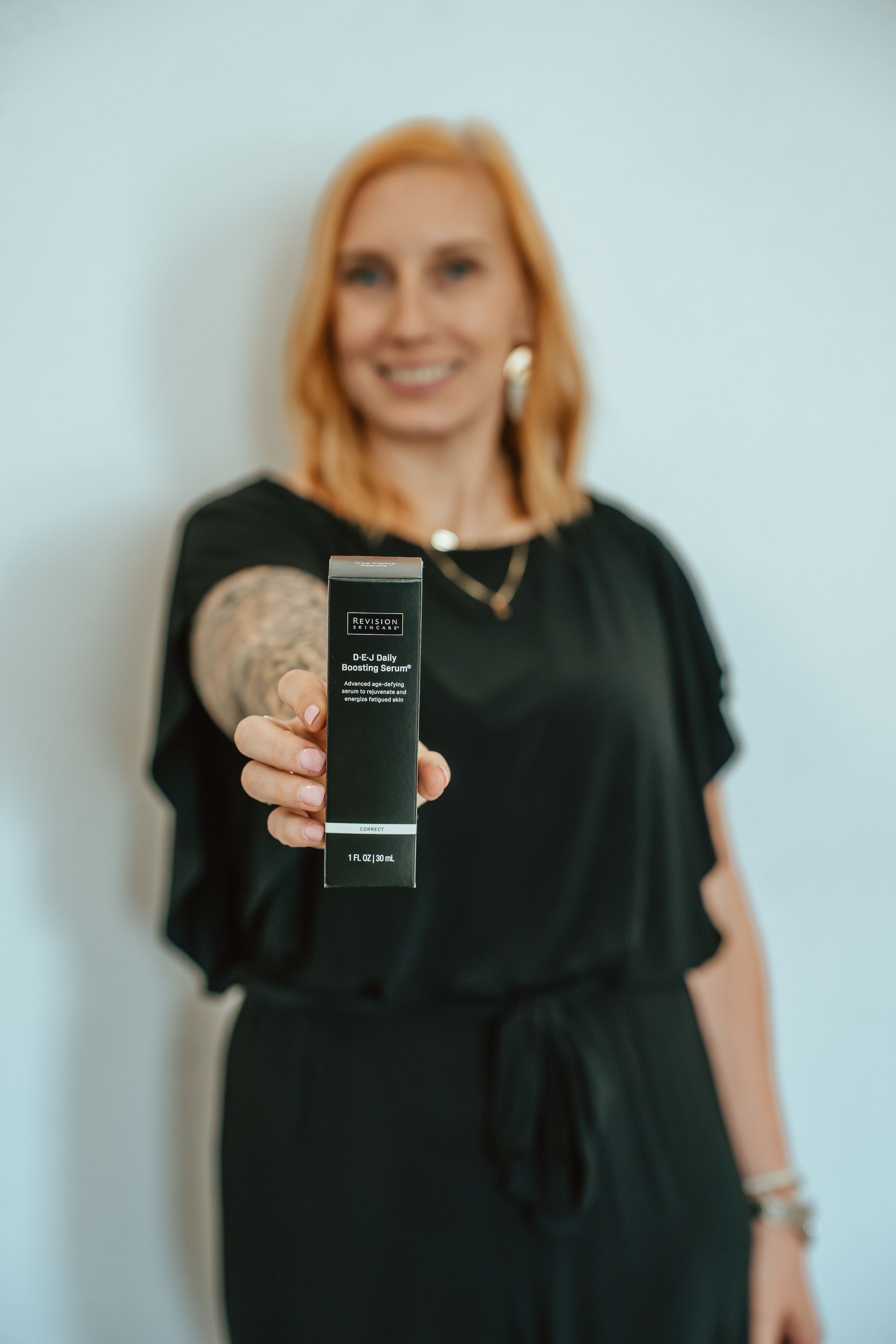 Woman in black dress holds a black product box, smiling at the camera.