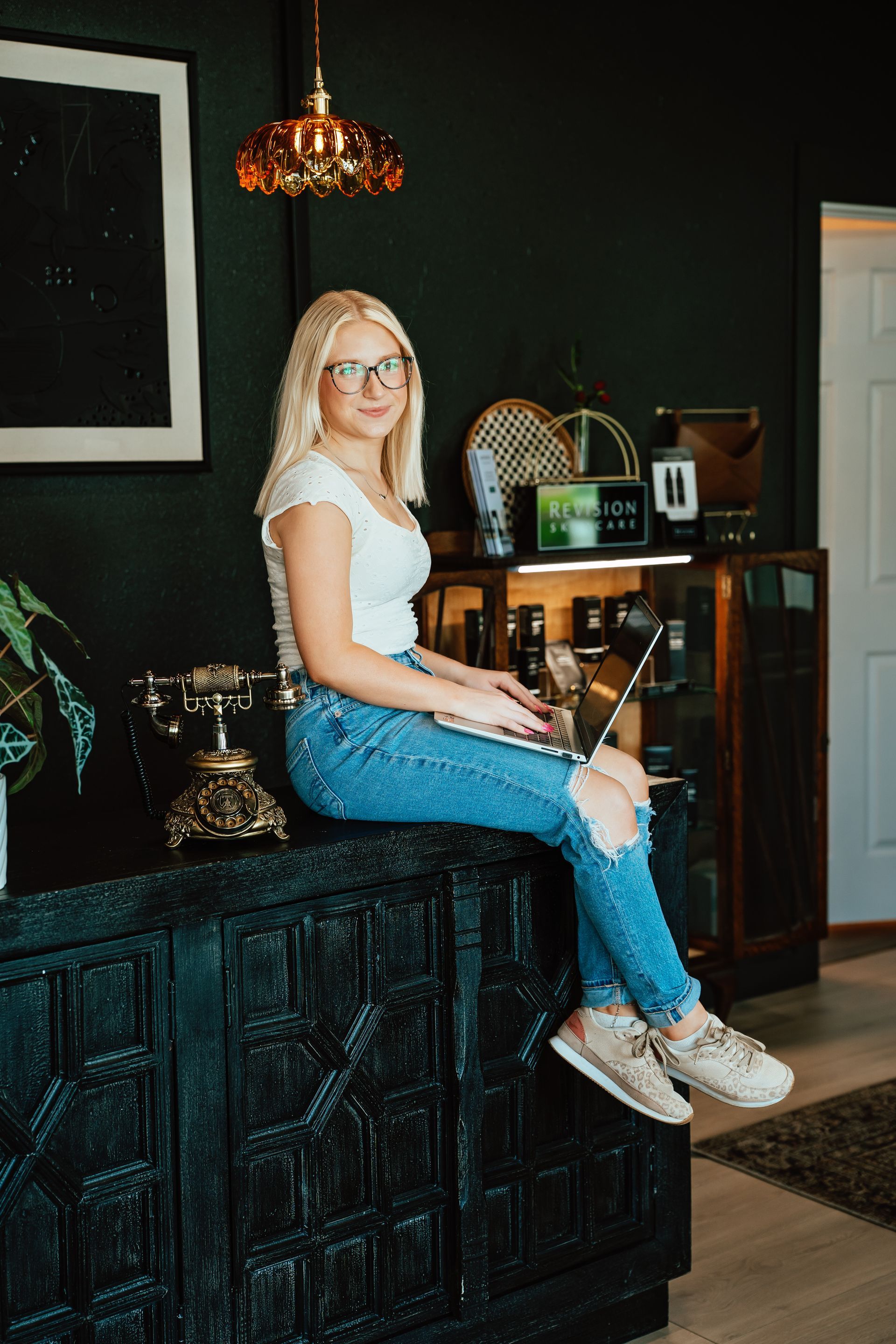 Blonde woman in glasses, ripped jeans, and white top sits on a black cabinet, working on a laptop.