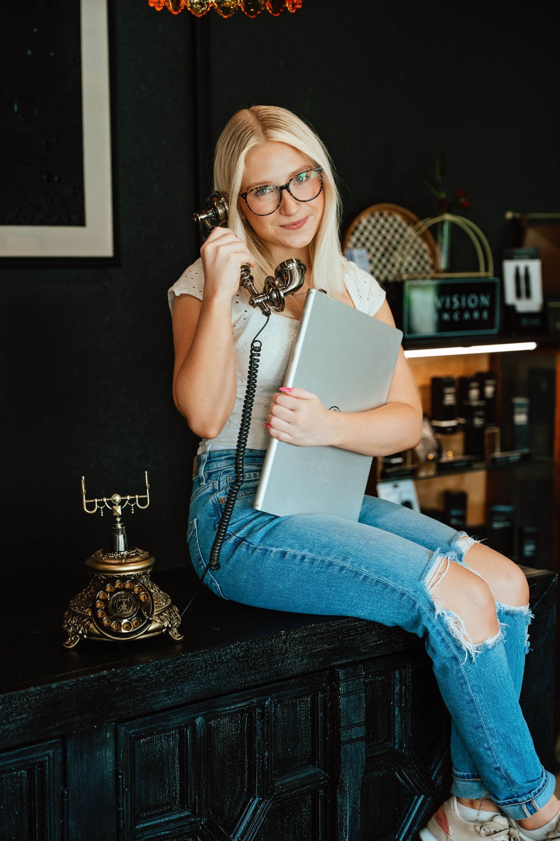 Blonde woman with glasses, holding laptop and antique phone, smiling. Sitting on a black table, indoors.