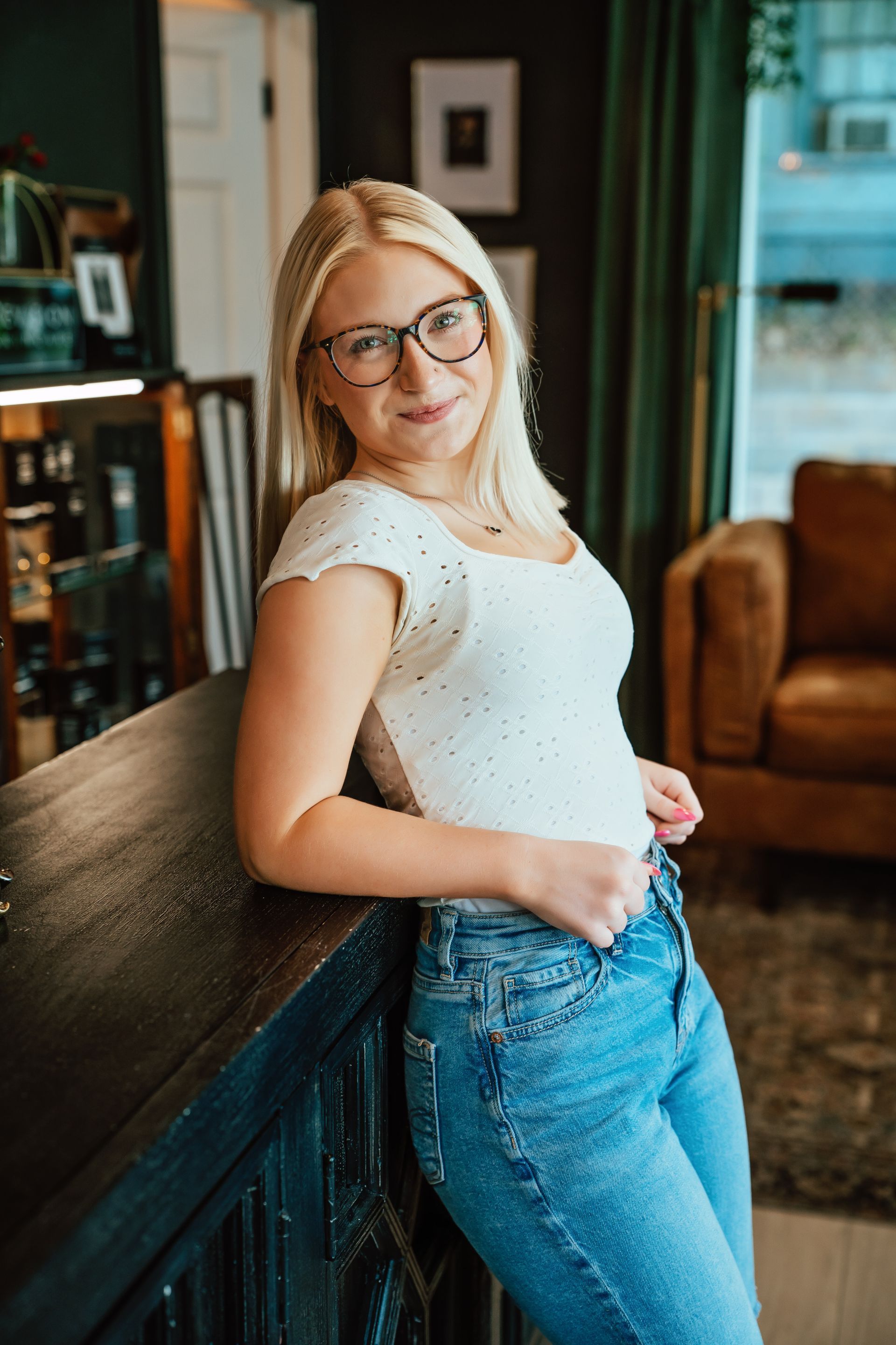 Blonde woman with glasses, wearing a white top and jeans, leans on a dark staircase.