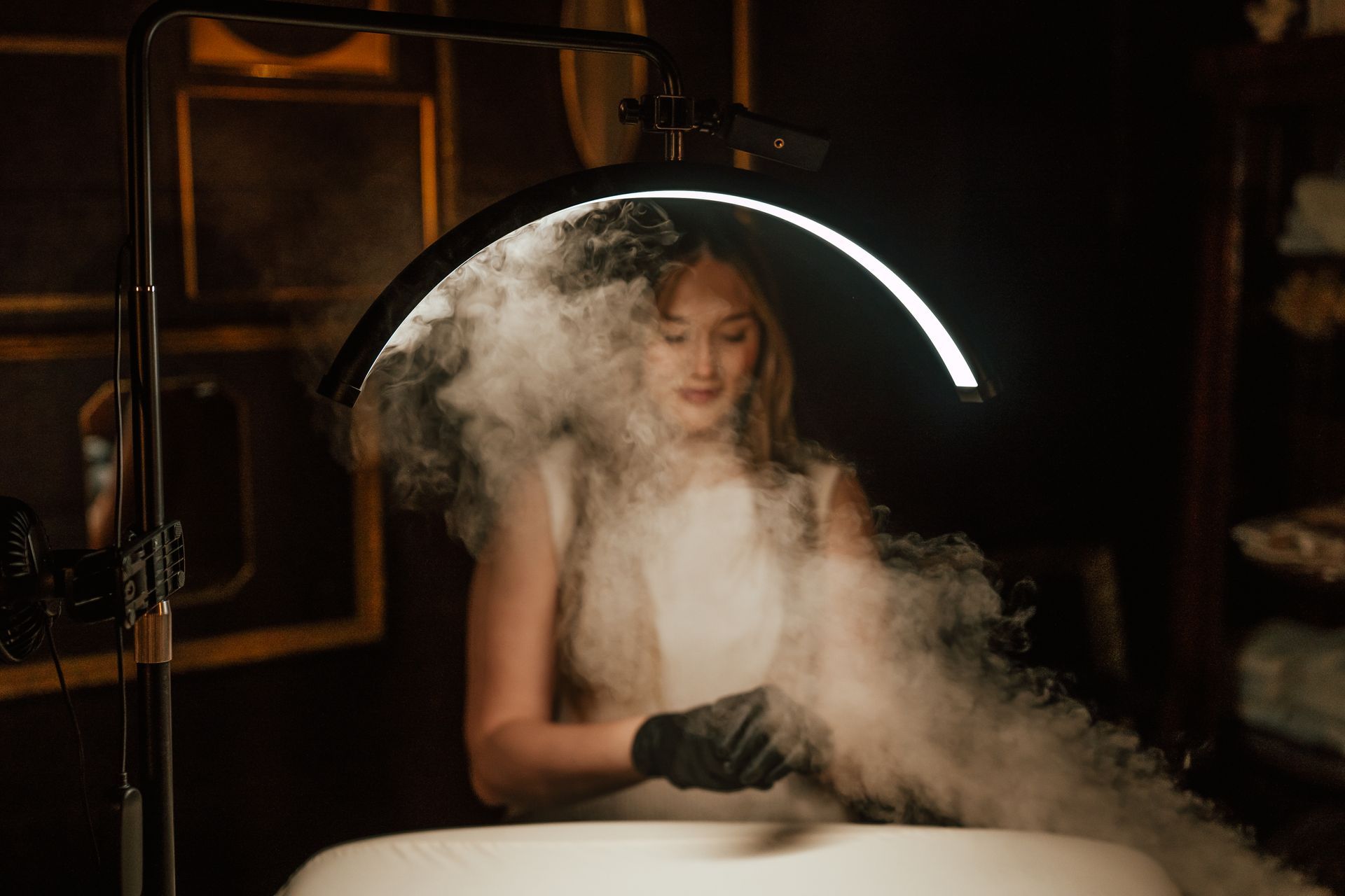 Woman in white dress and black gloves amid smoke under a bright ring light, indoors.