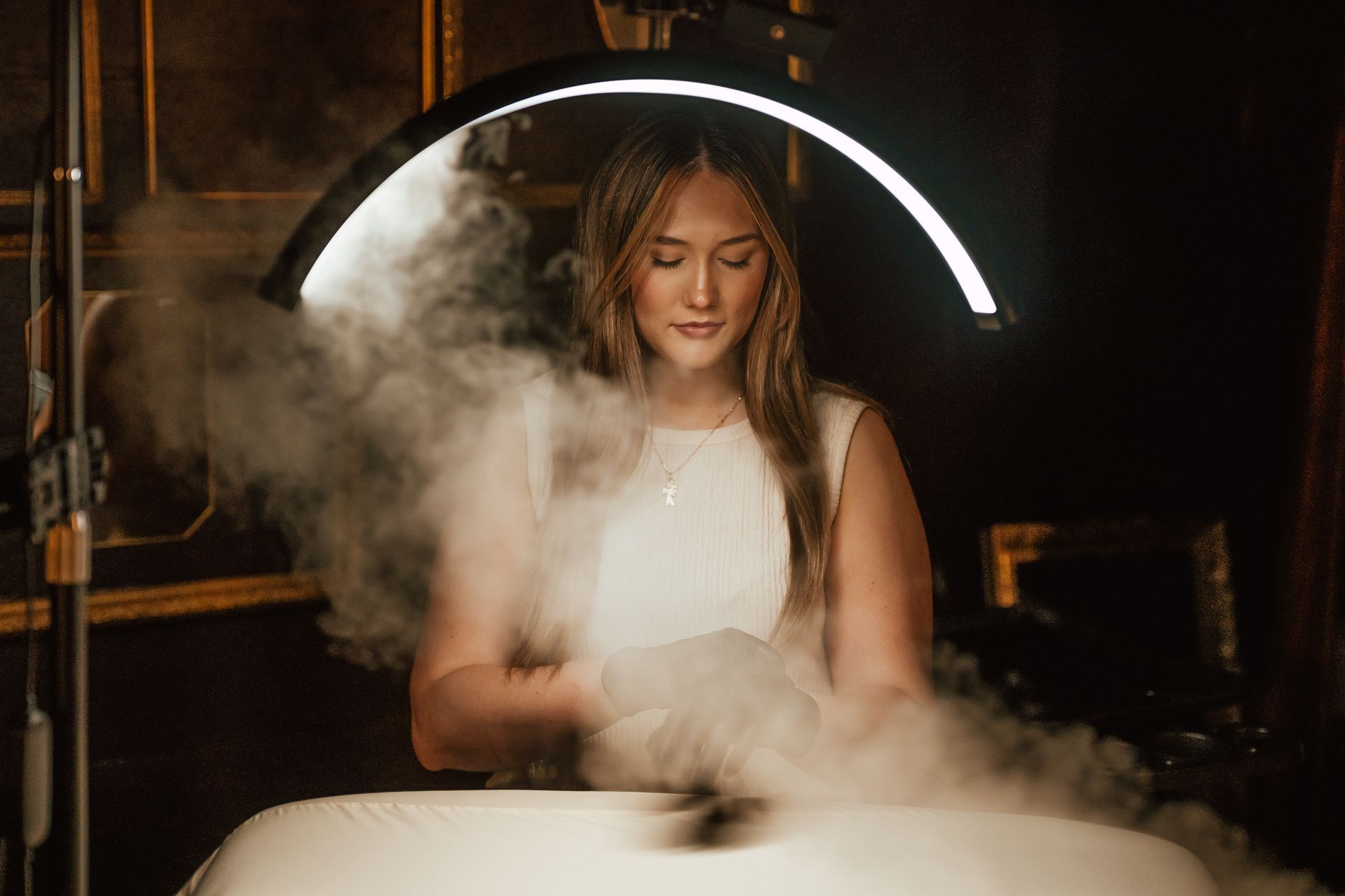 Woman in white dress surrounded by fog, under a bright ring light in a dark room.