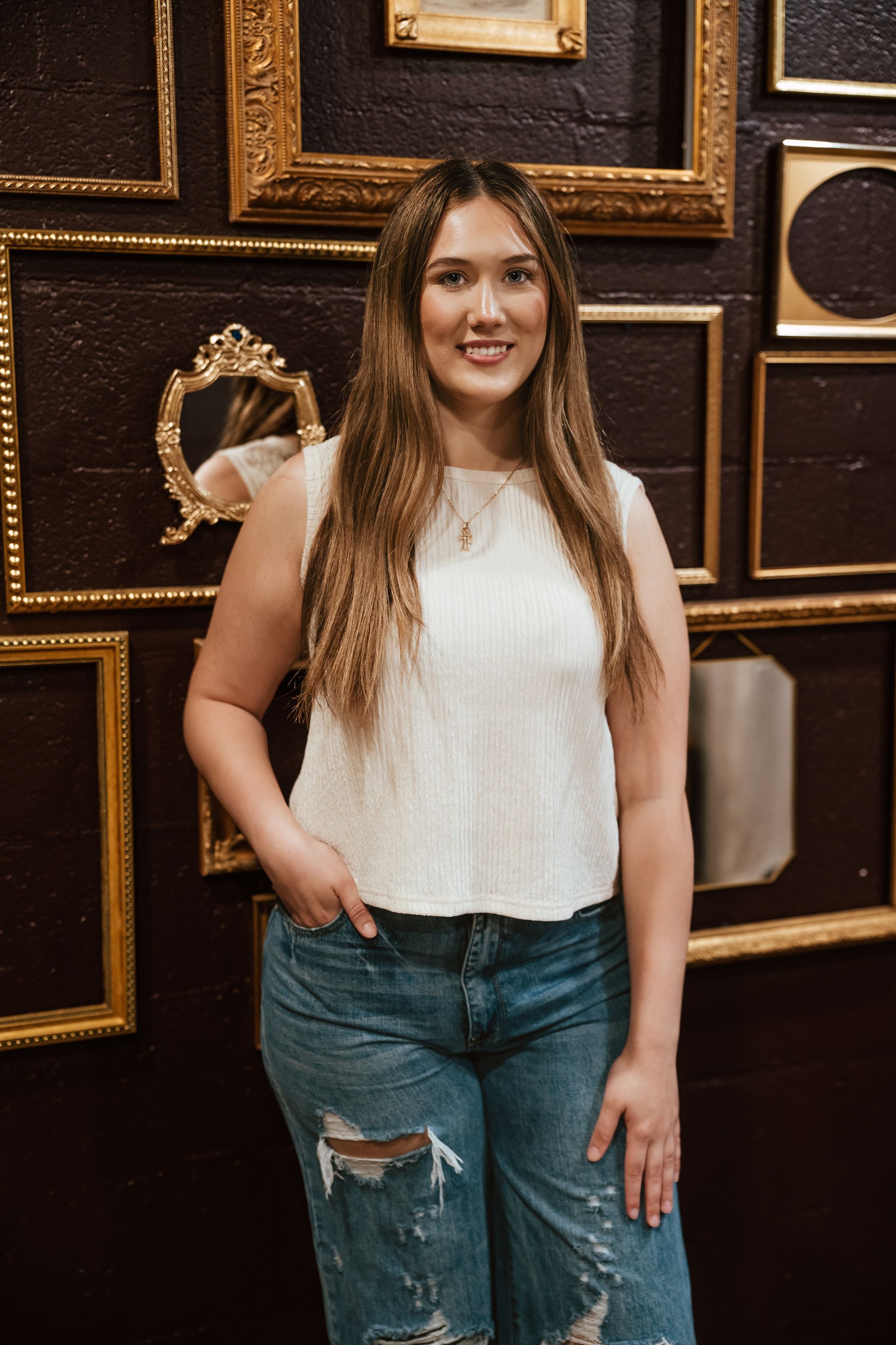 Woman in white top and ripped jeans smiles, posing in front of a wall with empty gold frames.