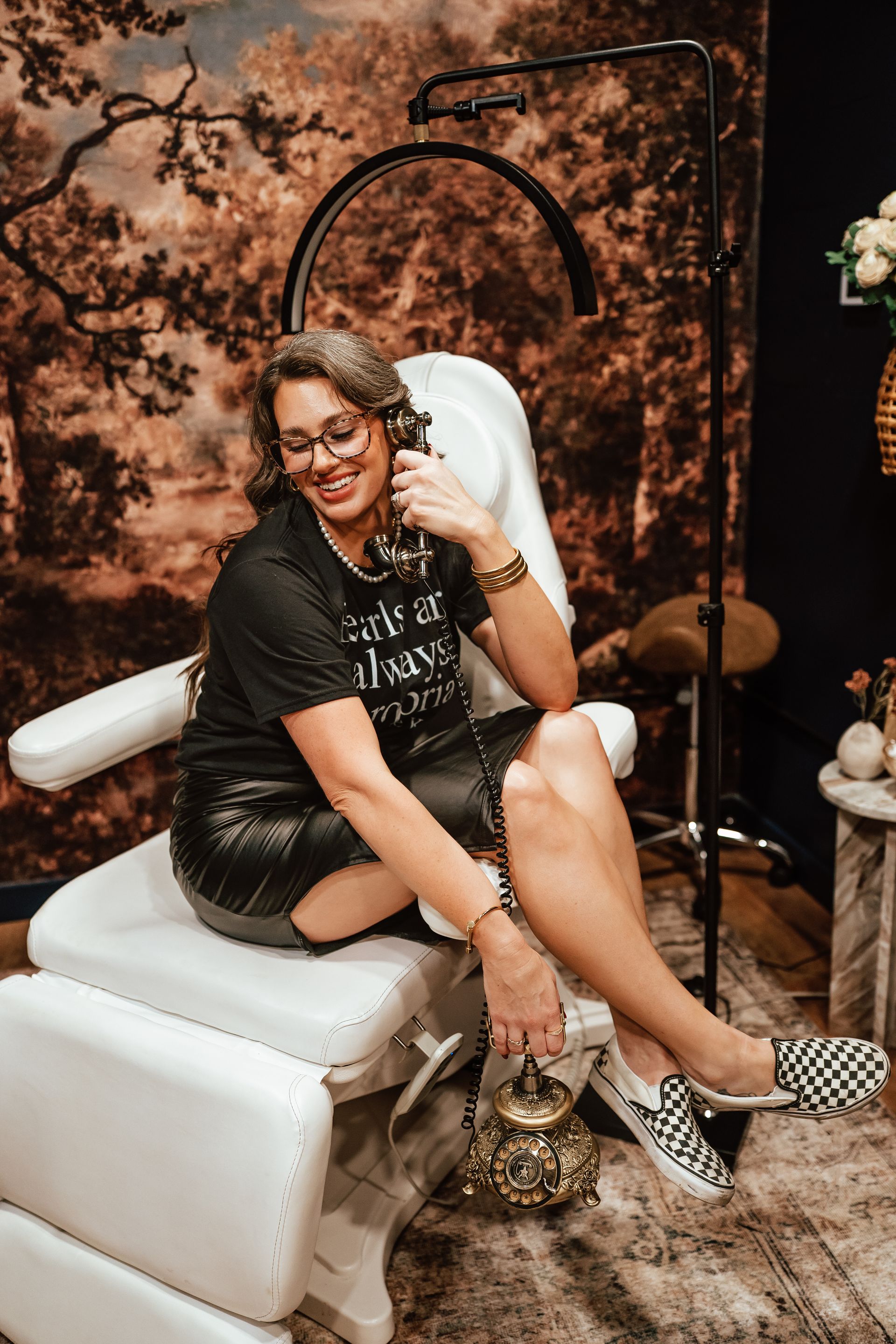 Woman in black shirt and leather skirt sitting in a white chair. Wearing glasses and checkered shoes.