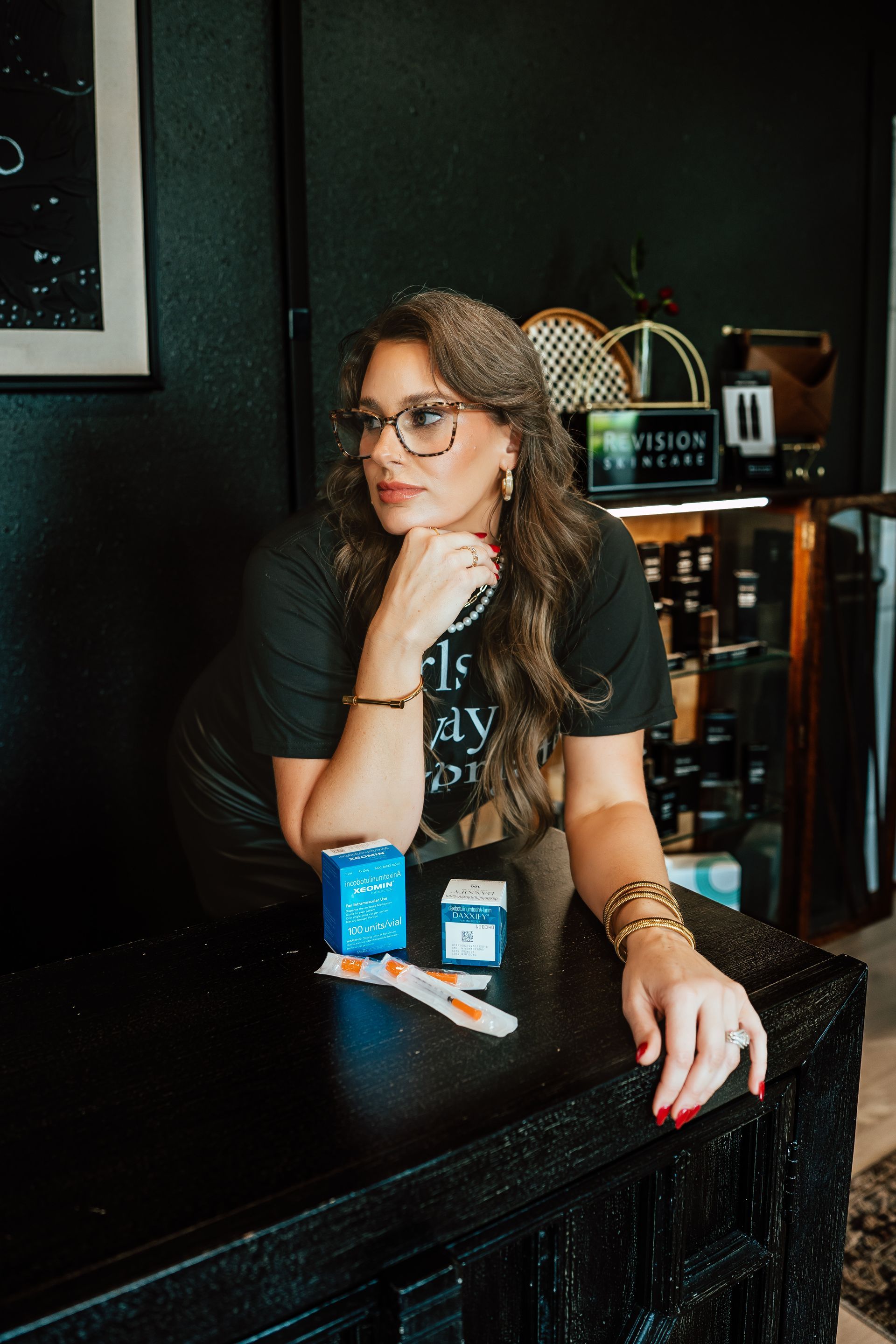 Woman in glasses, black shirt, gold jewelry, leans on a black table with product boxes.