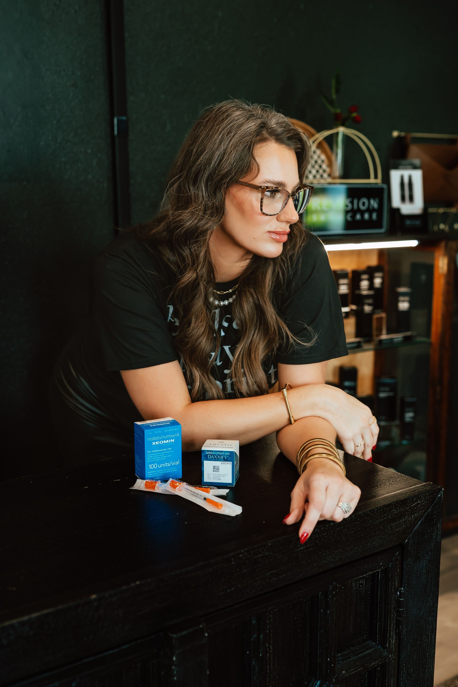 Woman with glasses at a counter, leaning on it, with two product boxes and a small brush. Dark interior.