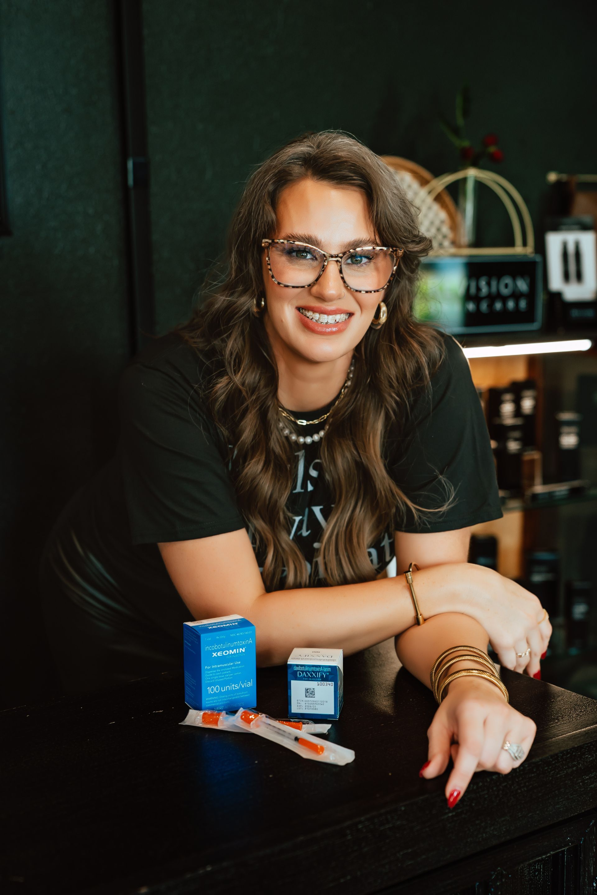 Woman with glasses leaning on a counter, displaying products. Dark setting, jewelry, and a smile.