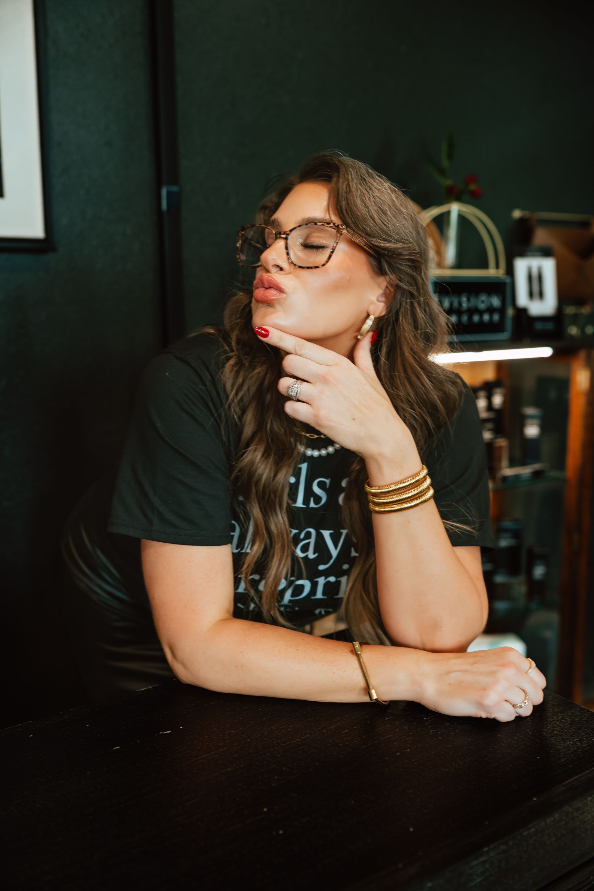Woman with glasses pouting, hand on chin, wearing black t-shirt, gold jewelry, in a shop setting.