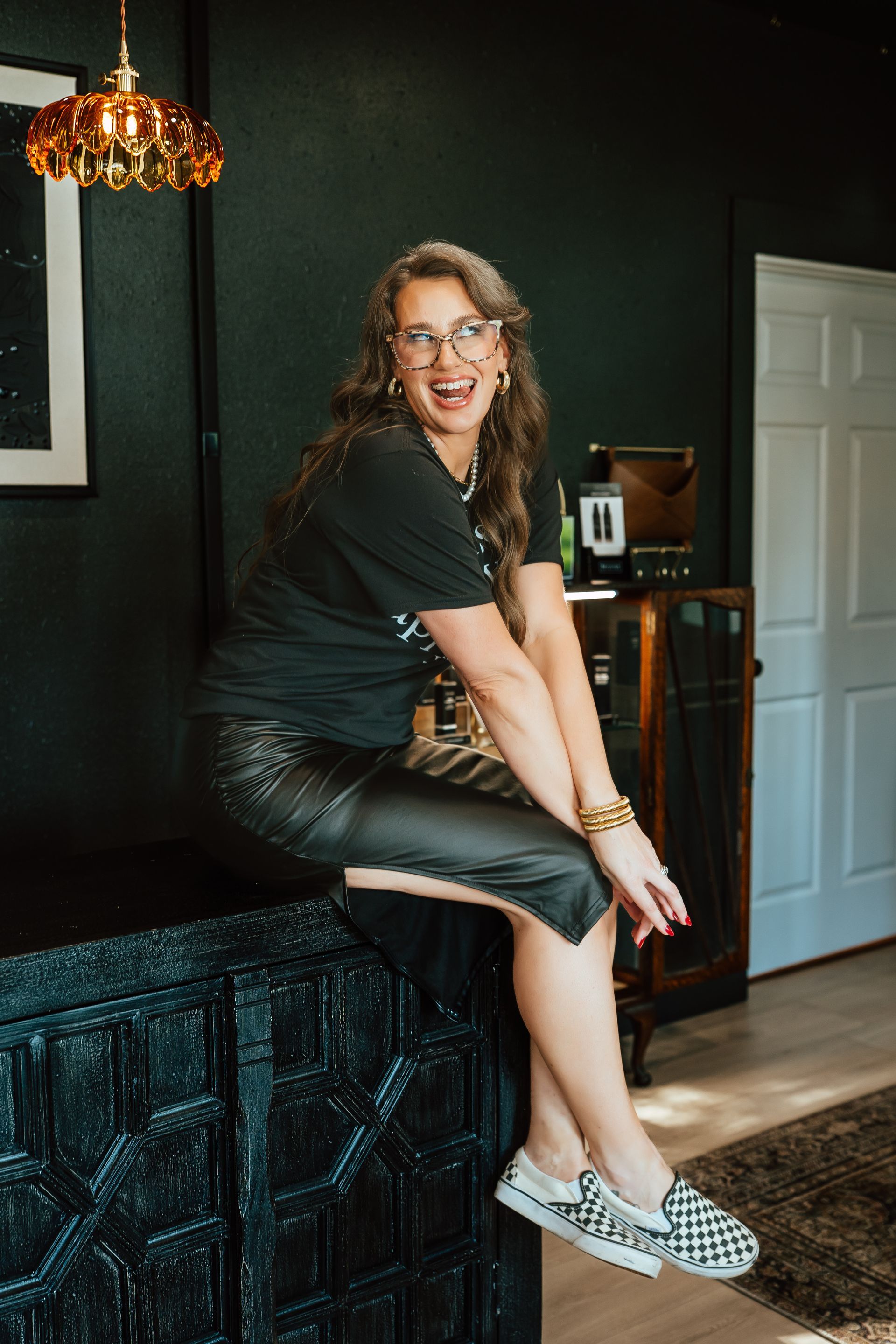 Woman in black leather skirt and checkered shoes, sitting on a dark dresser, smiling, indoors.