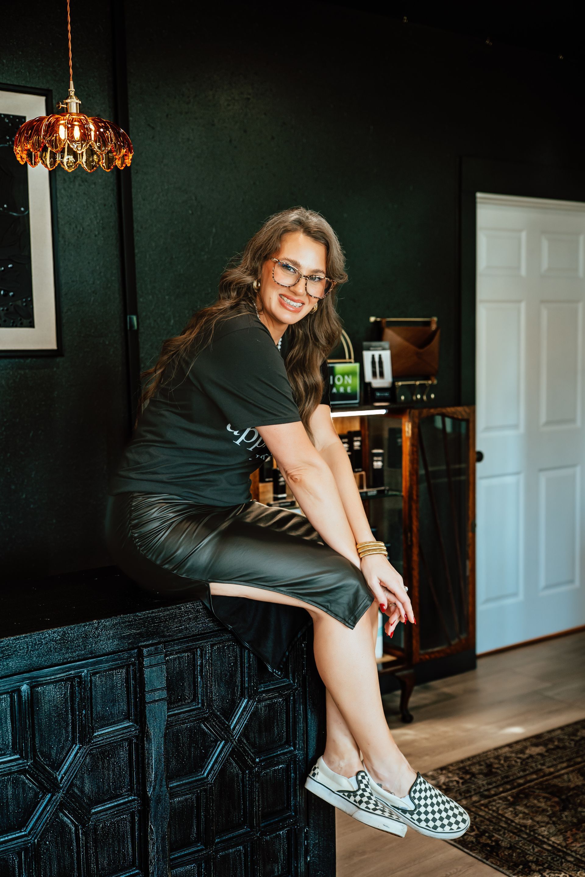 Woman in glasses sits on dark cabinet, smiling. Black wall, gold light fixture, and patterned shoes are visible.