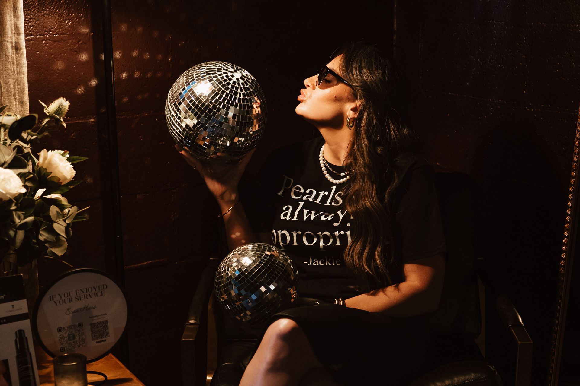 Woman kissing a disco ball in a dimly lit room. She wears sunglasses and a shirt saying 