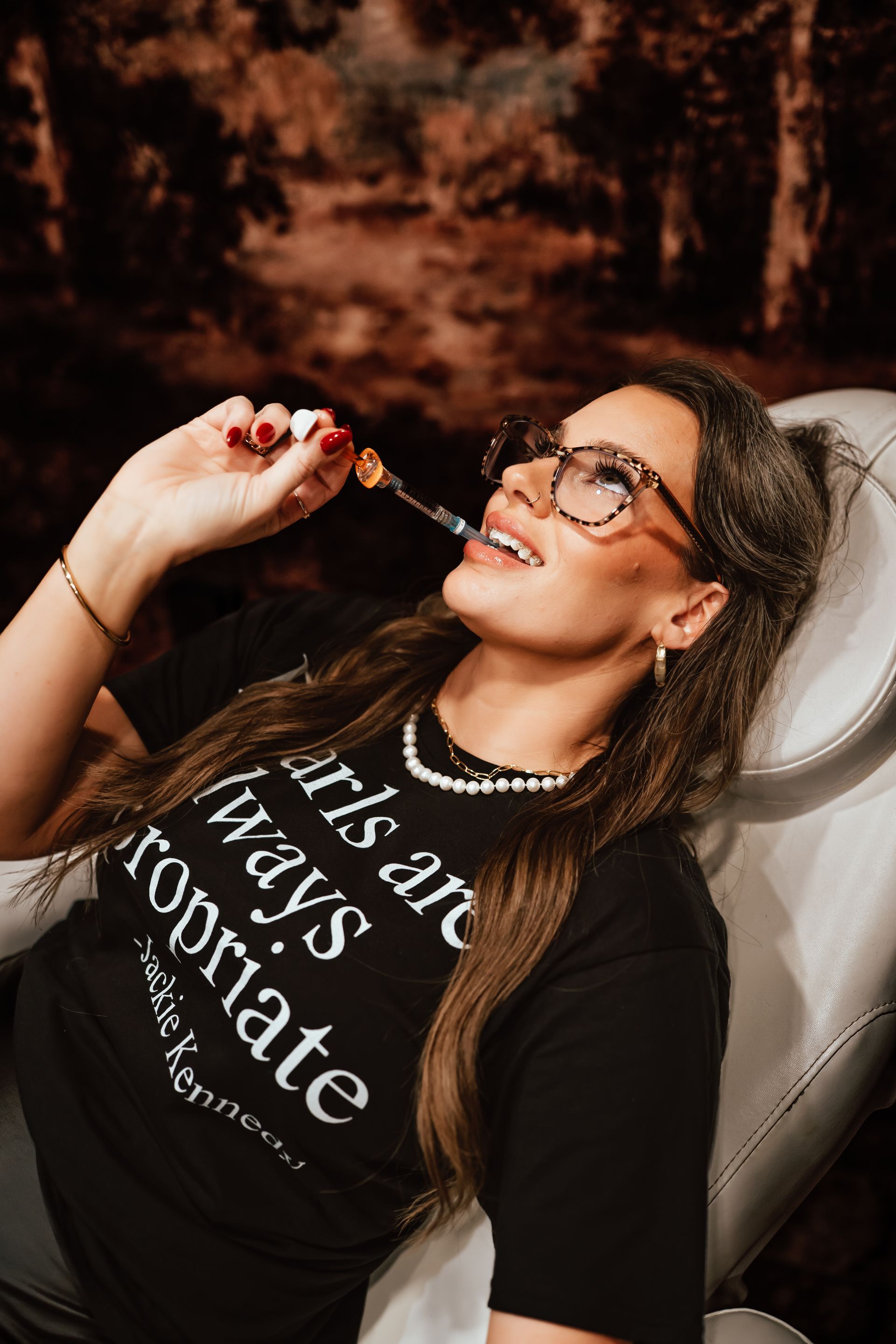 Woman in glasses, black shirt, smiling, holding a lollipop, in a dentist chair.