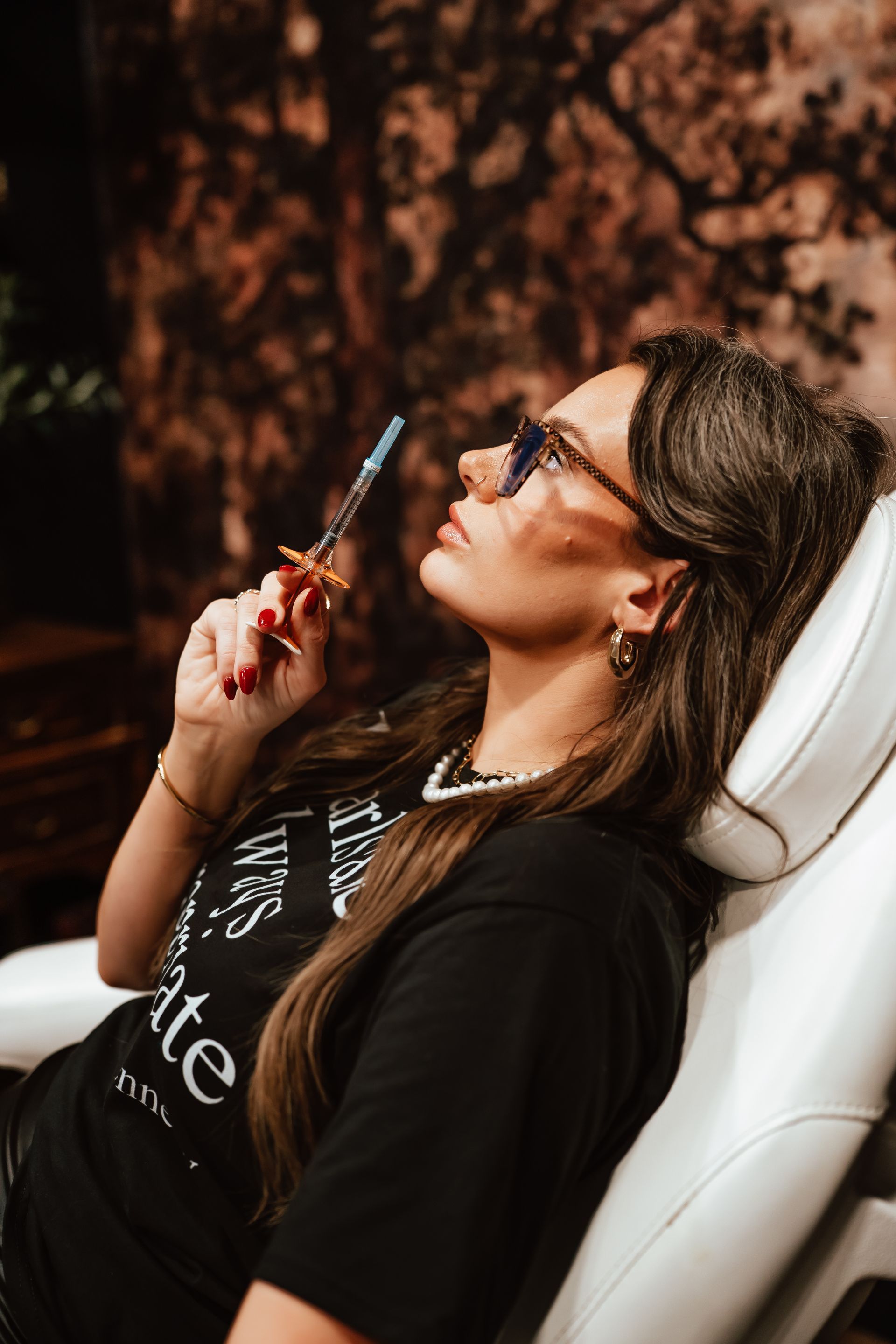 Woman in sunglasses holding a syringe, leaning back in a chair, looking up.