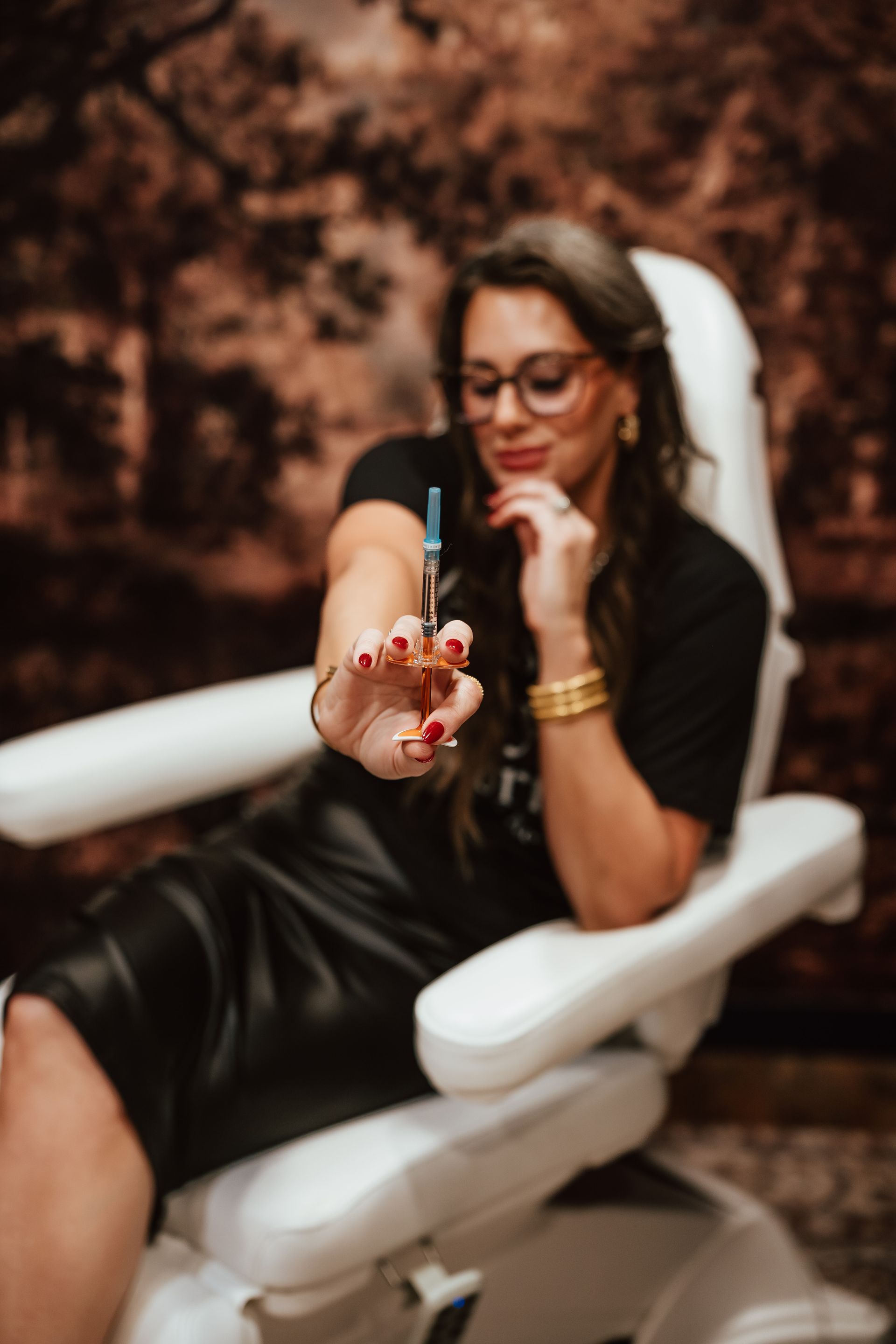 Woman in chair holding syringe, wearing glasses and jewelry, in a beauty treatment room.