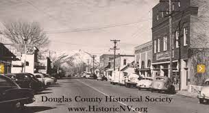 A black and white photo of a small town street with cars parked on the side of the road.