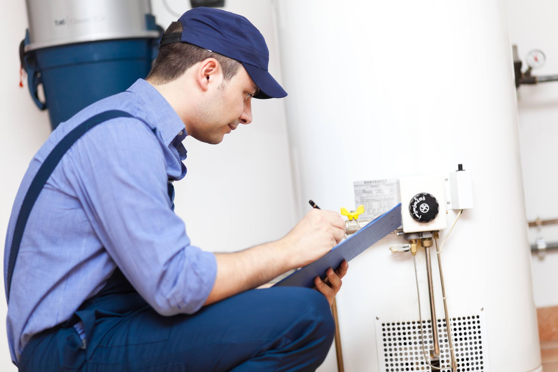 Plumber in blue uniform examines a water heater, writing on a clipboard in a utility room.