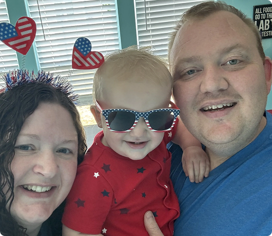 Family of three smiling, child wearing American flag sunglasses and shirt. Patriotic decorations in background.
