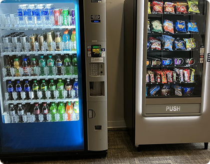 Two vending machines: one for drinks, blue-lit, and one for snacks, gray. Located in a room.