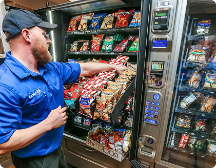 Man in a blue shirt restocking a vending machine with various snacks, indoors.