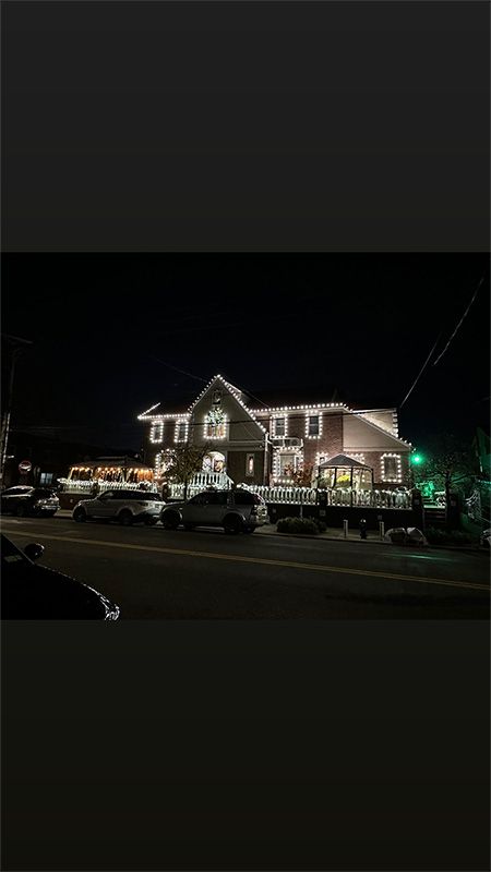 A large house at night, lit with white lights along the roofline. Cars parked on the street in front.