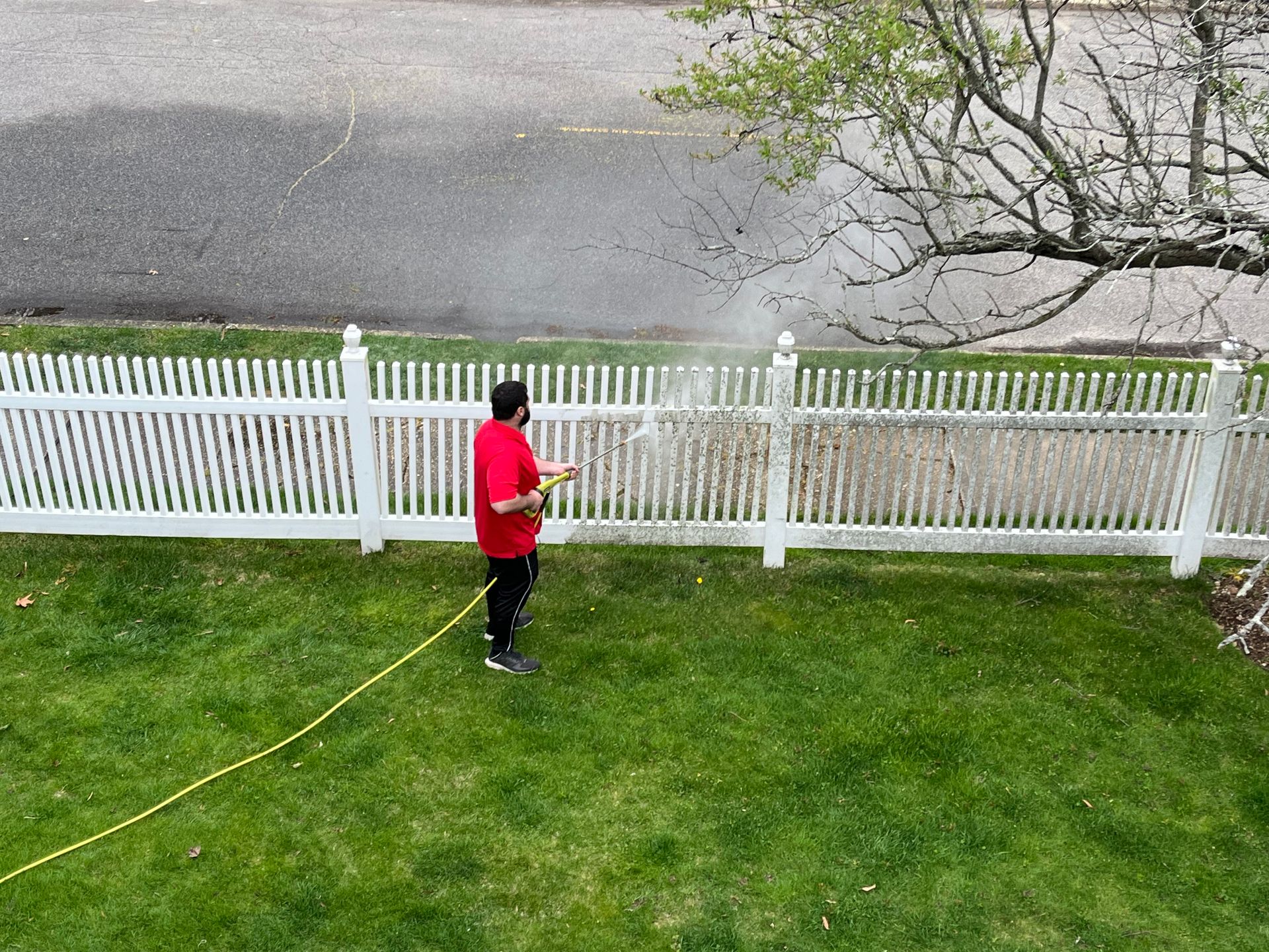 A man in a red shirt is spraying water on a white picket fence.