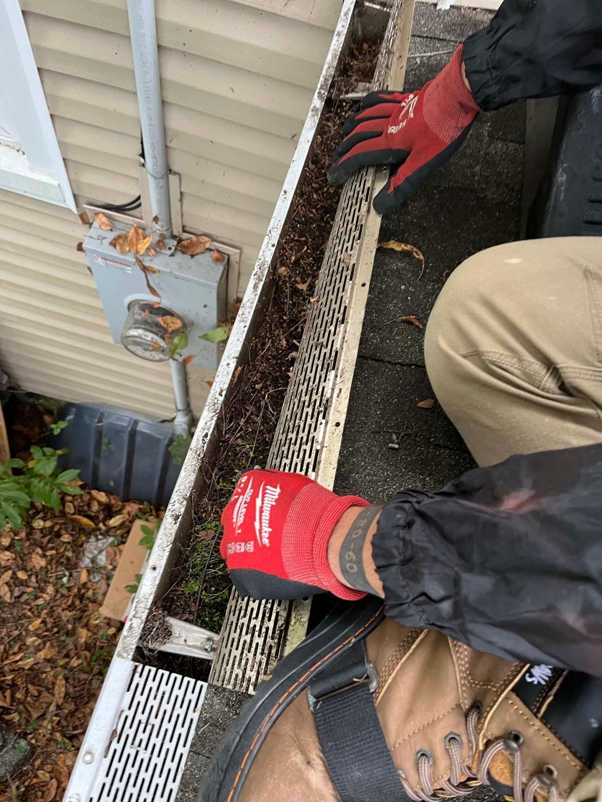 A person wearing red gloves is cleaning a gutter on the side of a house.
