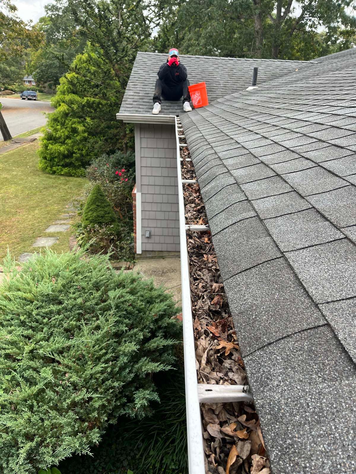 A man is sitting on the roof of a house cleaning a gutter.
