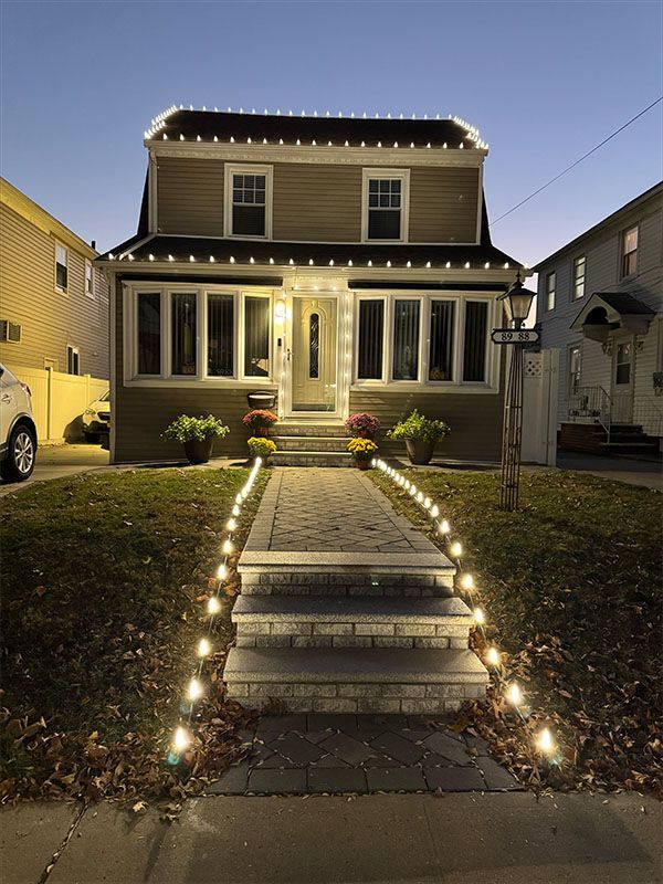 Two-story house at dusk, lit with white lights along the roofline, walkway, and steps, with flowers on the porch.