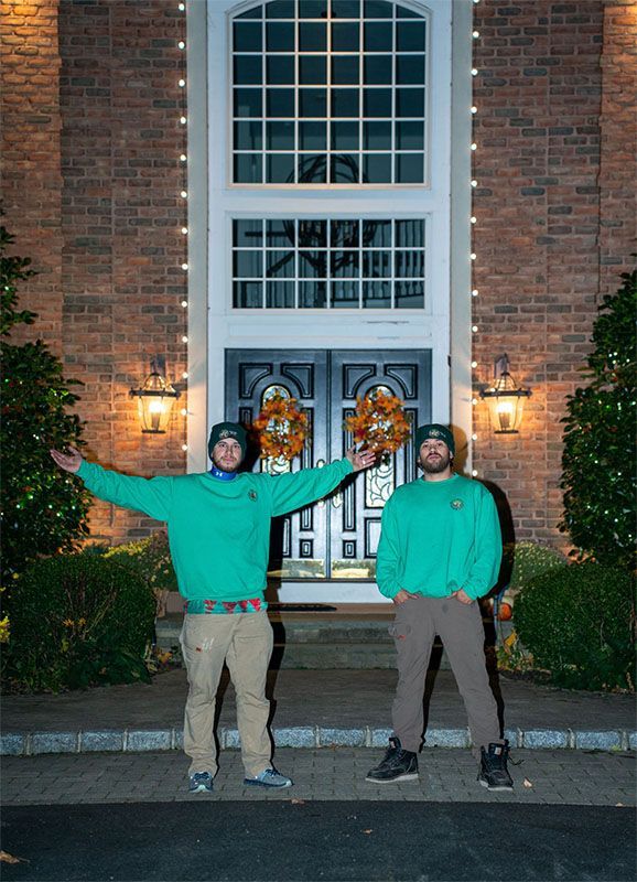 Two men in green sweaters and hats pose in front of a brick house with fall wreaths on the doors.