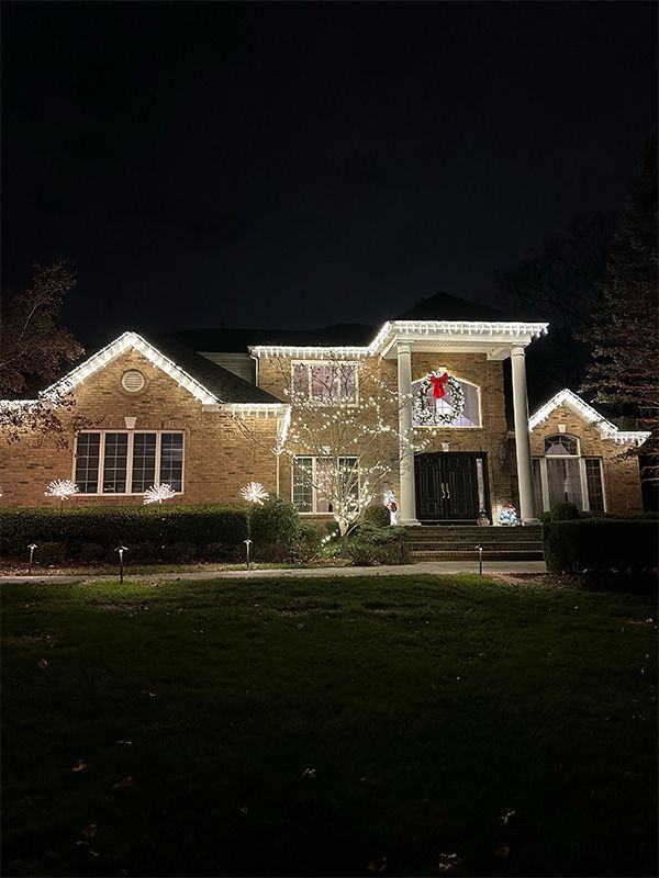 A brick house at night, illuminated with white Christmas lights. A wreath hangs over the front door.