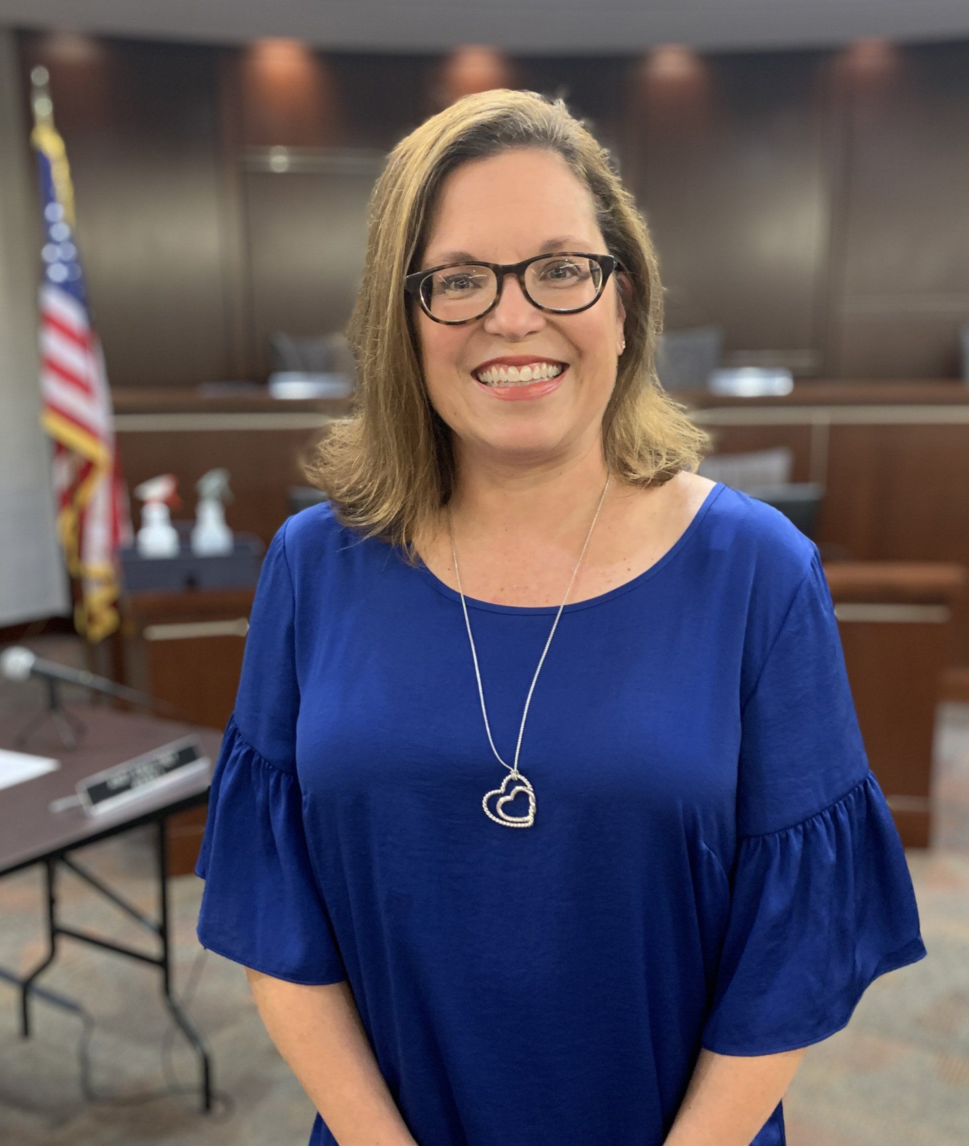 A woman wearing glasses and a blue shirt is smiling in front of an american flag.